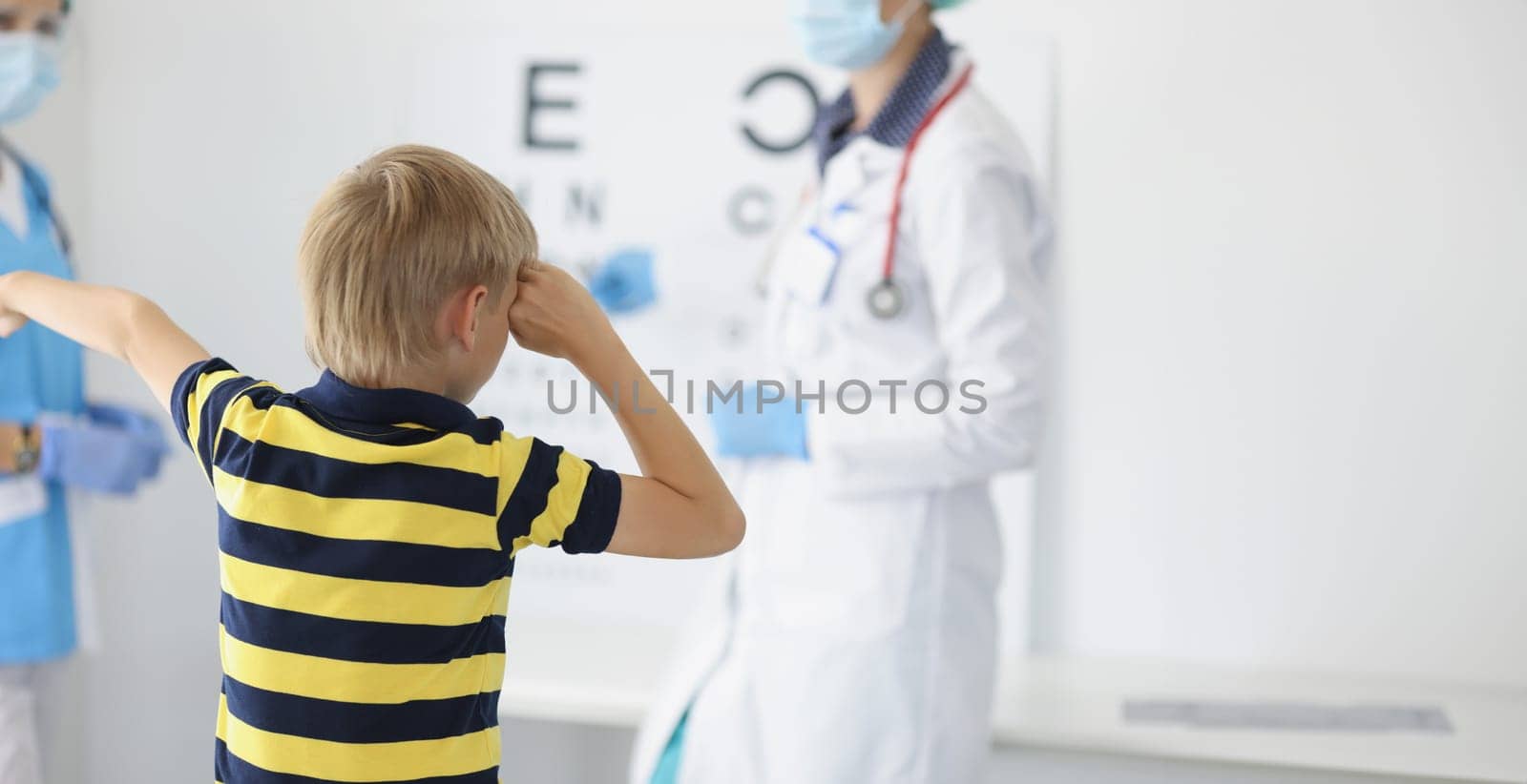 Portrait of boy on appointment in clinic, oculist cabinet, sight diagnostic closing eye and say out loud letter on board. Health, ophthalmology concept