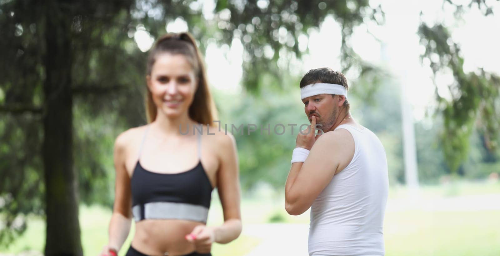 Portrait of middle aged man check on young beautiful fit woman running in park. Active woman jogging in front, feeling energetic. Sport, health concept