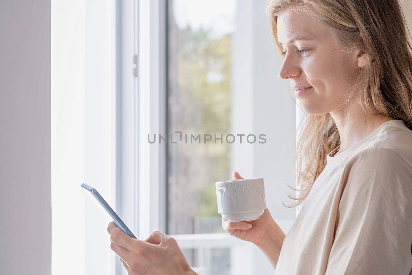 Woman standing beside the window using mobile phone holding coffee cup by Desperada