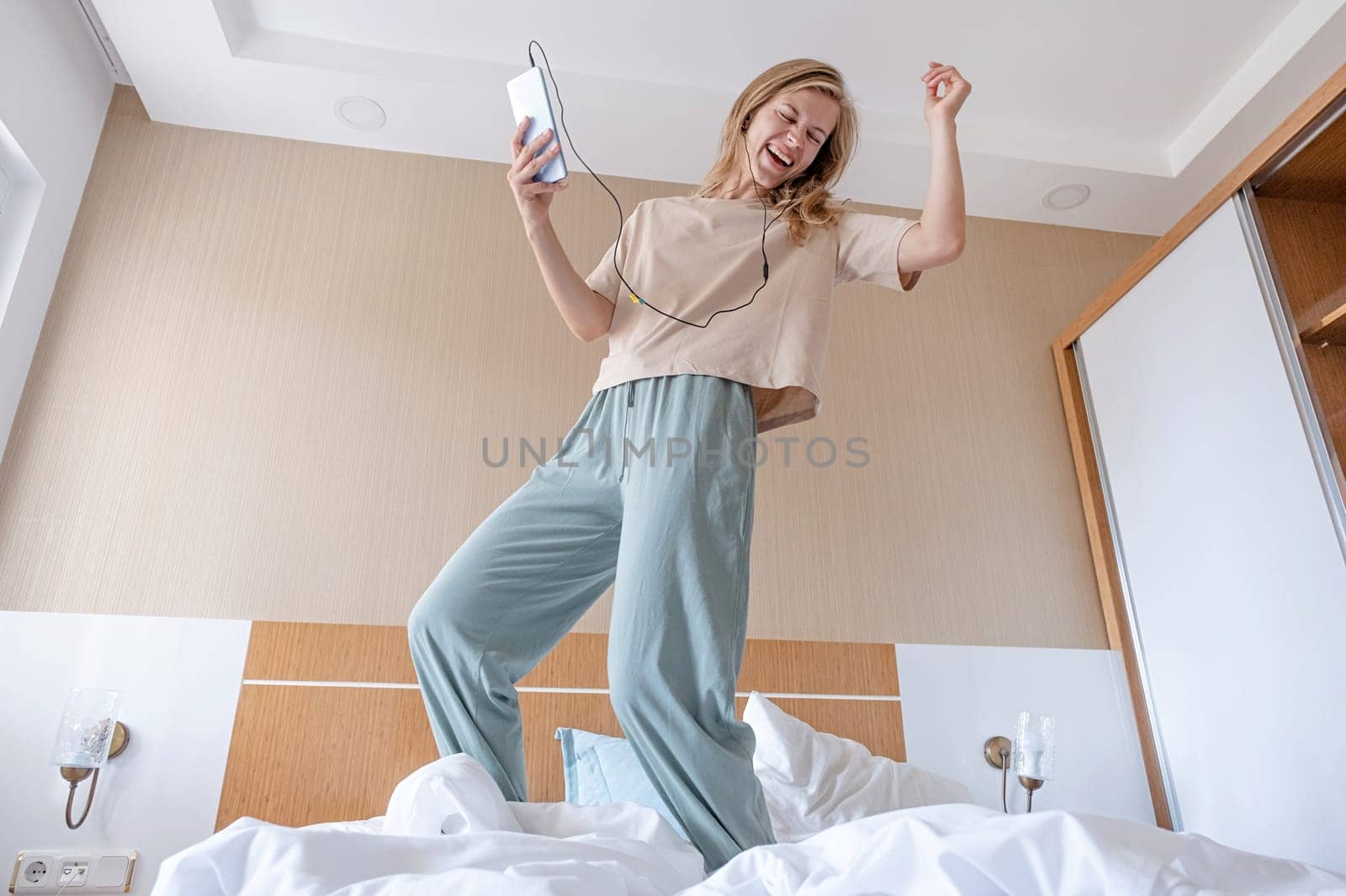 full length portrait of excited happy young female wearing pajama dancing to favorite music on bed in hotel room
