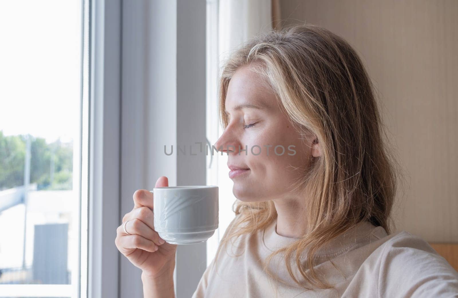 Happy woman standing by the window at hotel room drinking coffee by Desperada