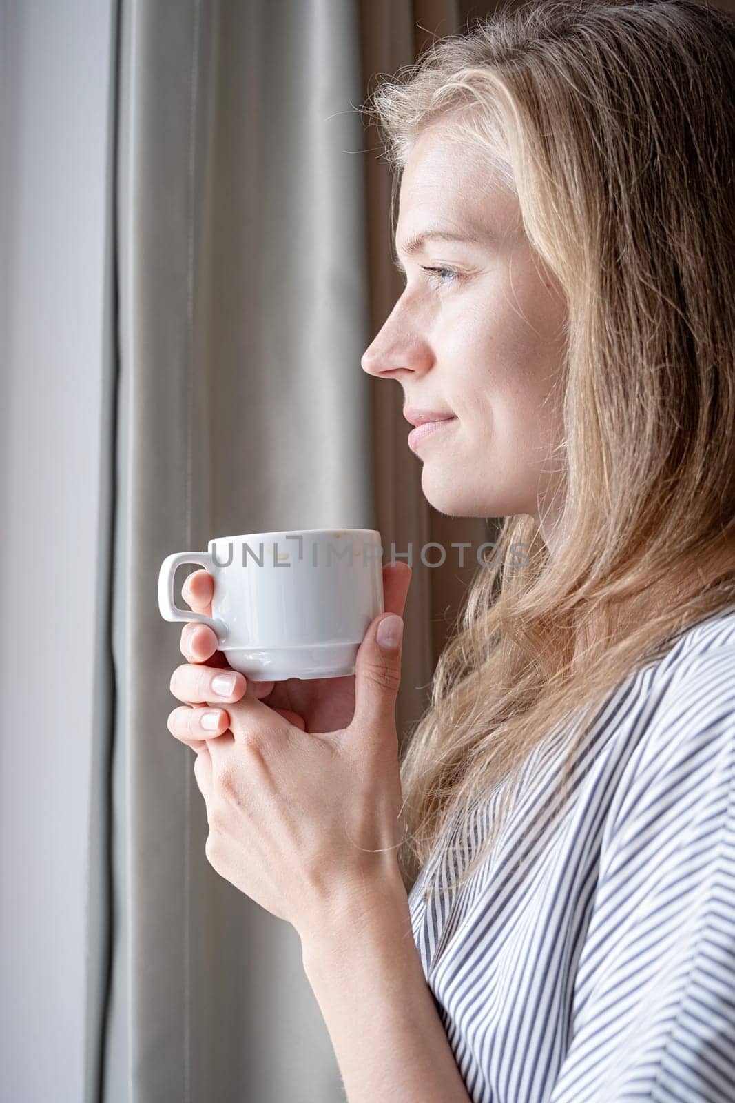 Happy woman standing by the window at hotel room drinking coffee by Desperada