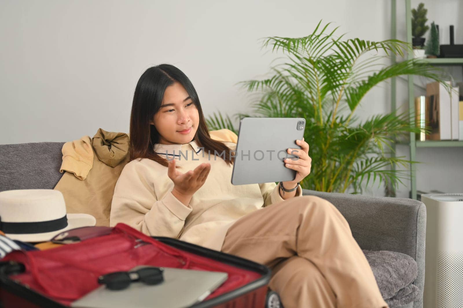 Smiling Asian woman having video call on digital tablet while preparing travel suitcase at home by prathanchorruangsak