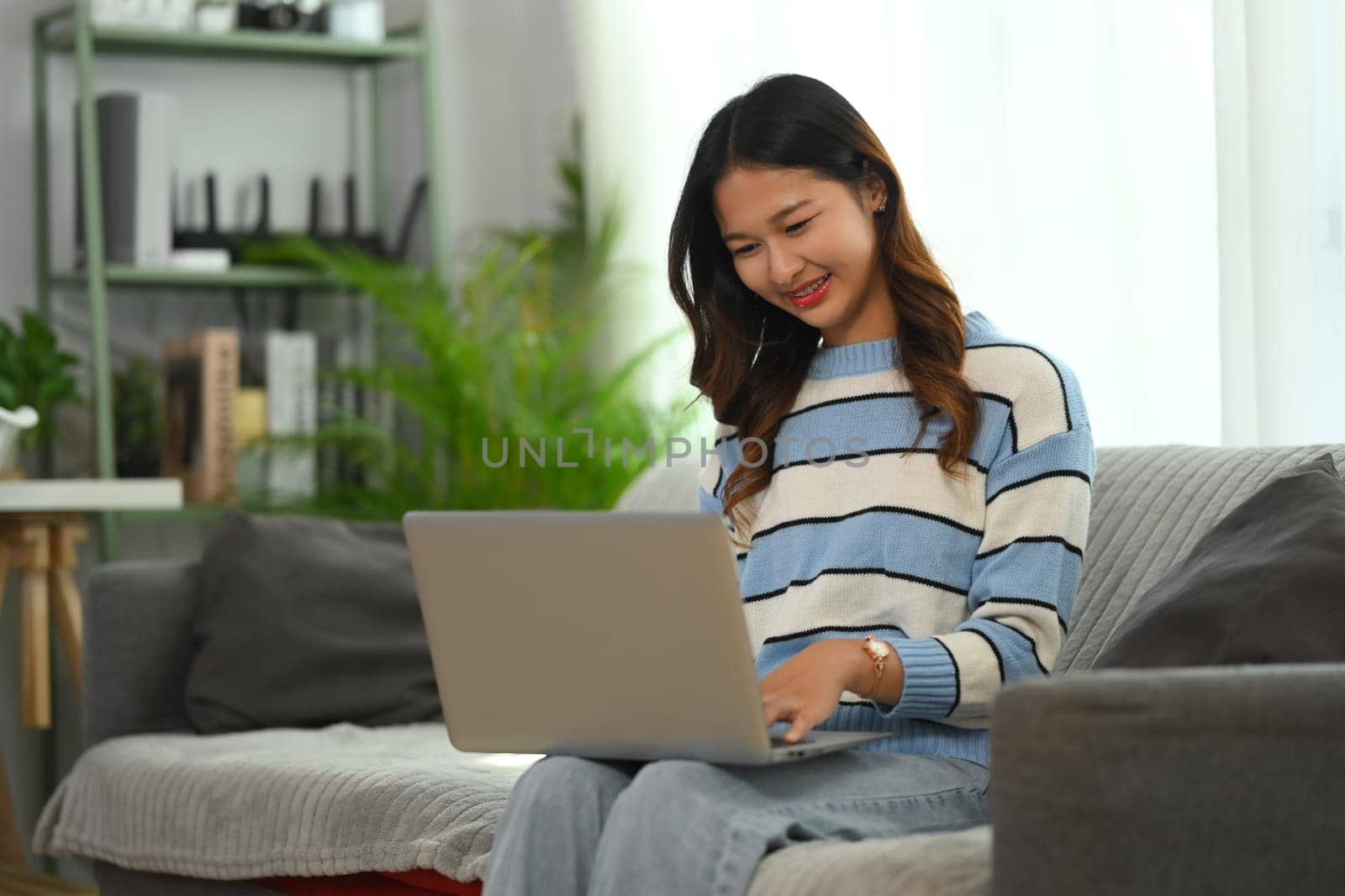 Smiling young woman relaxing on couch at home and using laptop. People and technology concept by prathanchorruangsak