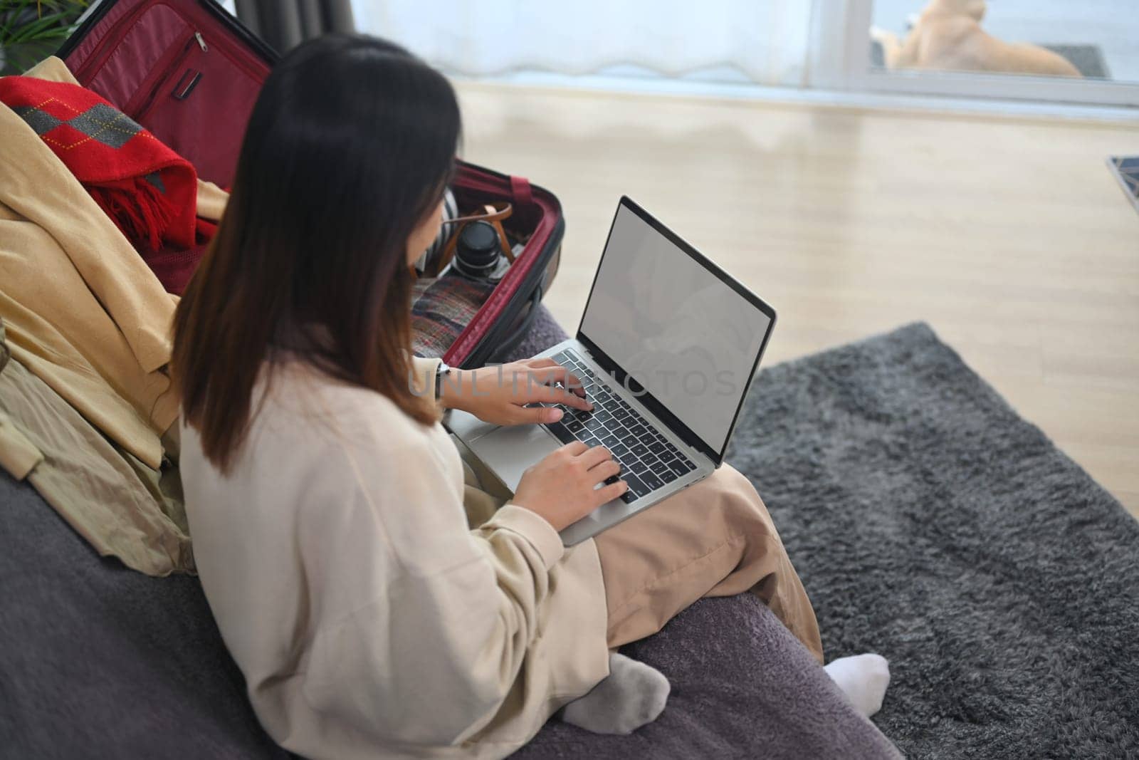 Young woman with travel bag sitting on couch using laptop for booking hotel. Travel and vacation concept.