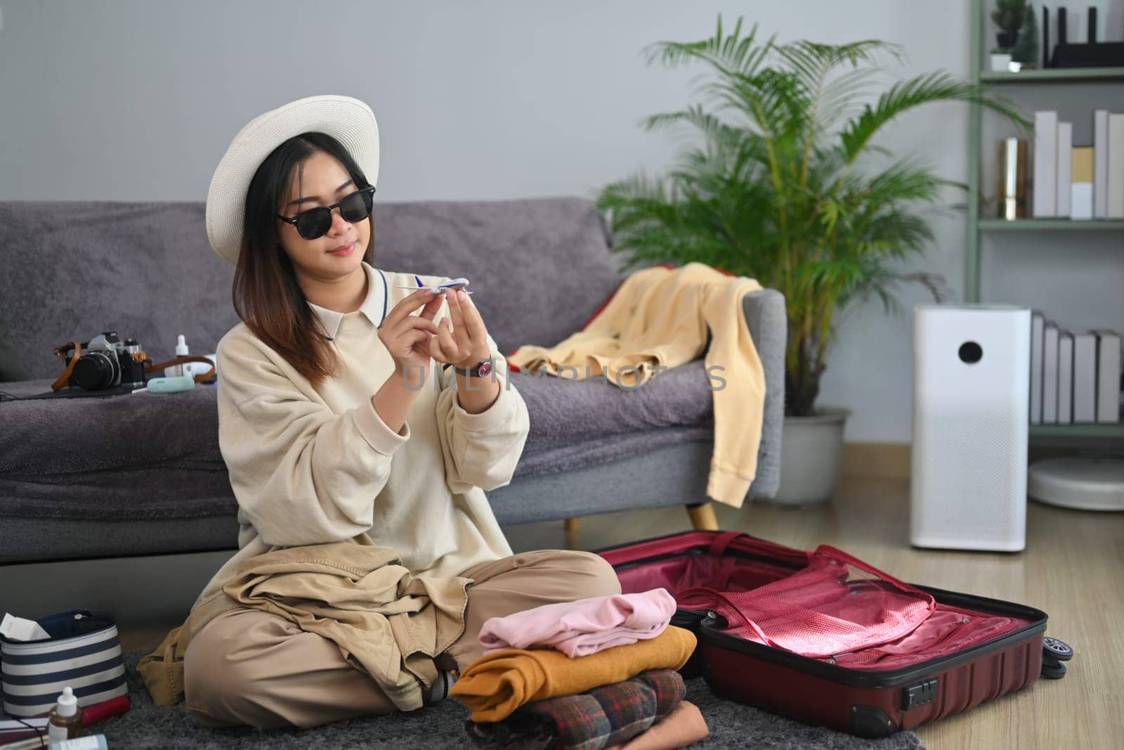 Happy woman traveler in sunglasses sitting on floor with suitcase hold airplane model in hands. Travel and vacation concept.