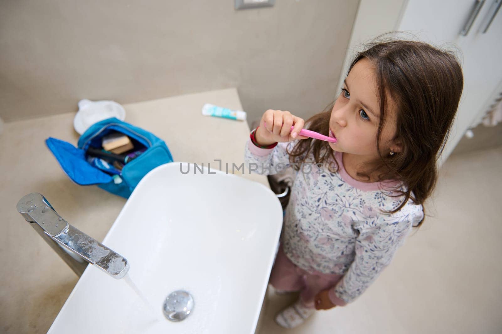 View from above of a child girl brushing teeth, standing at white washbasin in the home bathroom. Dental hygiene concept by artgf