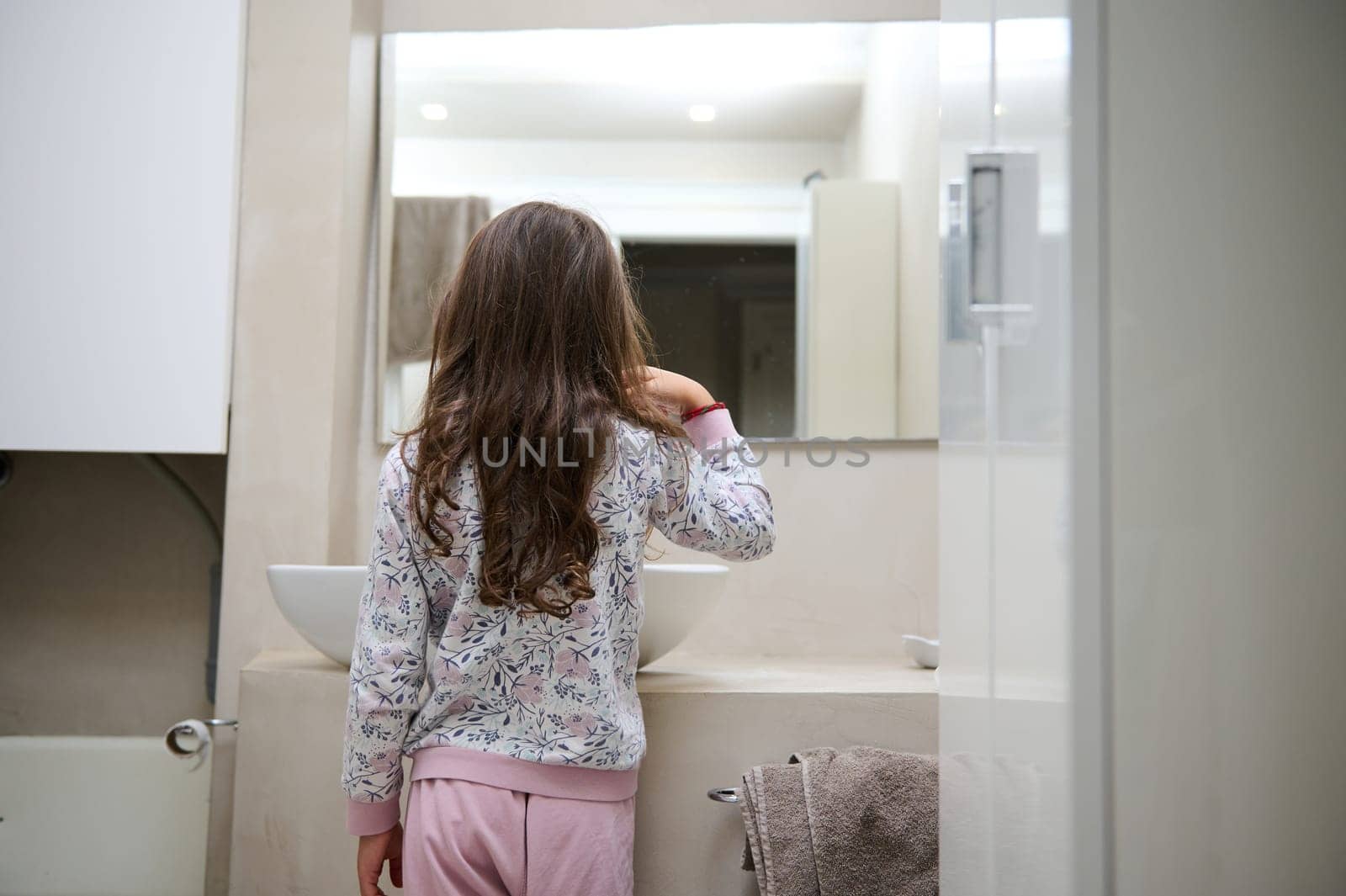Rear view child girl standing at washbasin in the home bathroom and brushing her teeth, looking at herself in the mirror by artgf