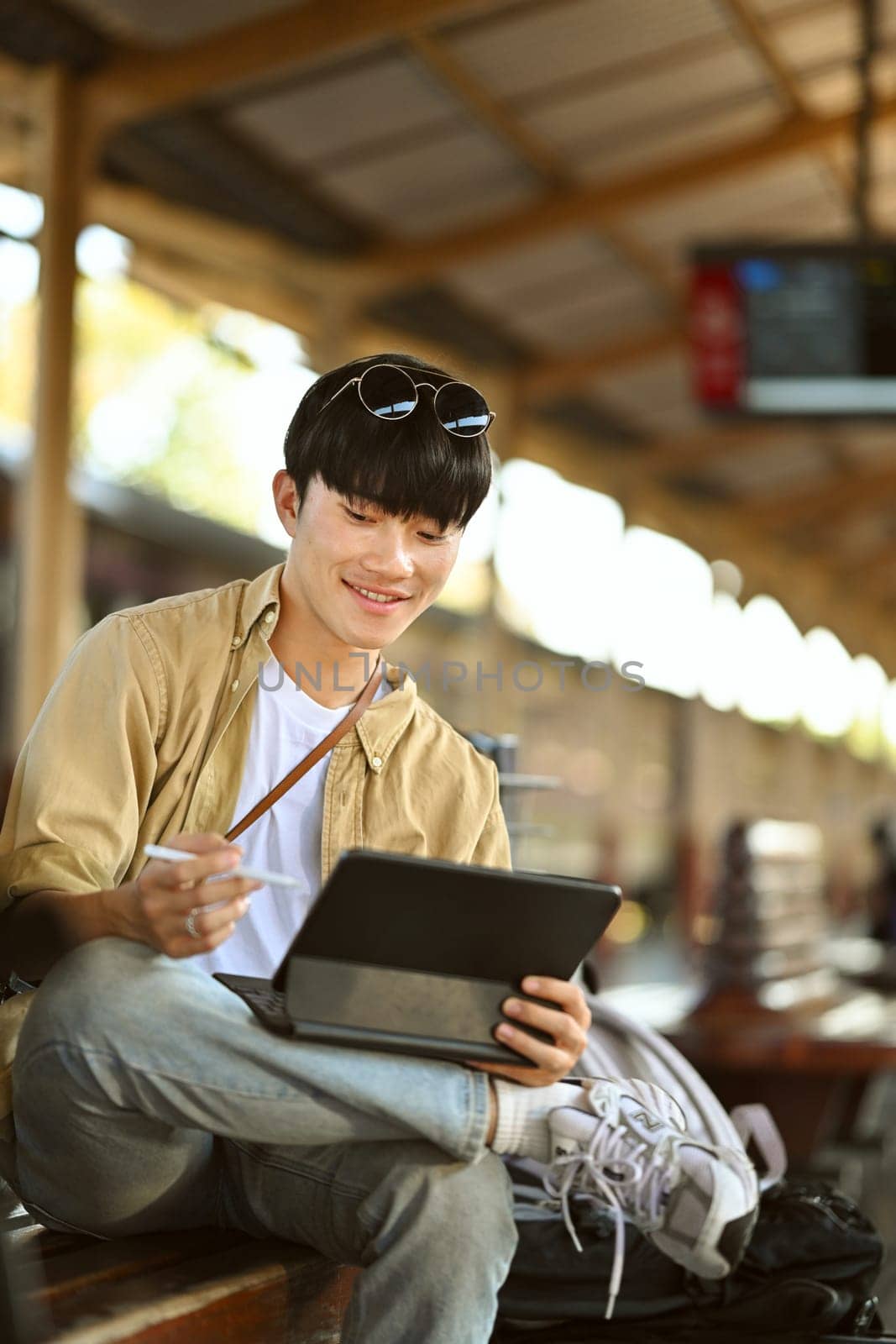 Portrait of young man with a backpack using digital tablet, sitting on a bench at the train station.