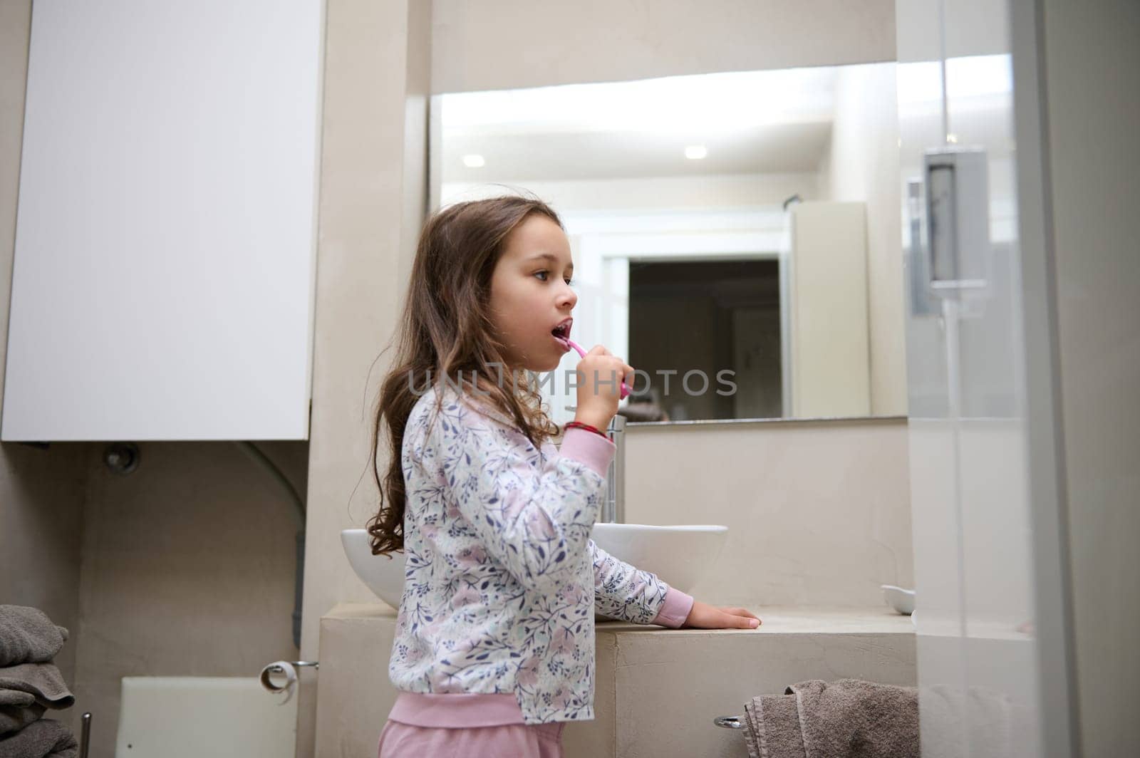 Side view of a Caucasian beautiful little kid girl girl in pajamas, brushing her teeth, while waking up in the morning, standing in the gray minimalist home bathroom. Dental hygiene and oral care.