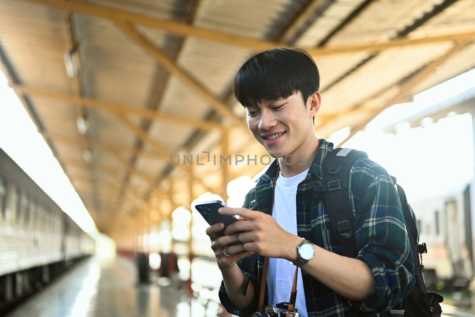 Smiling young man standing on the platform of a train station and using mobile phone.