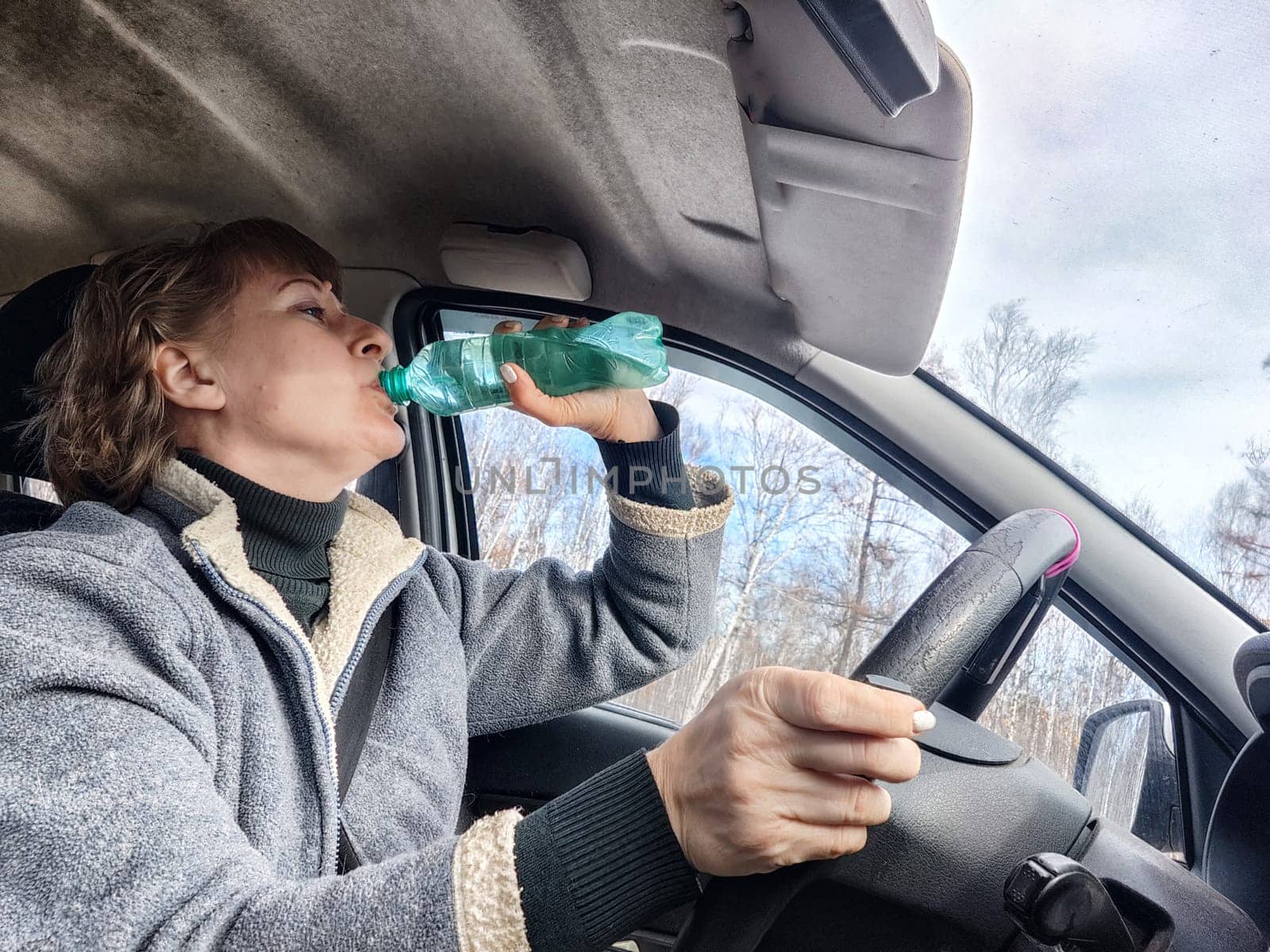 Portrait of female driver in solo journey. Adult mature woman holding steering wheel and bottle of water. Drinking while driving for to stay awake. Lady girl drinks alcohol vodka or gin while driving