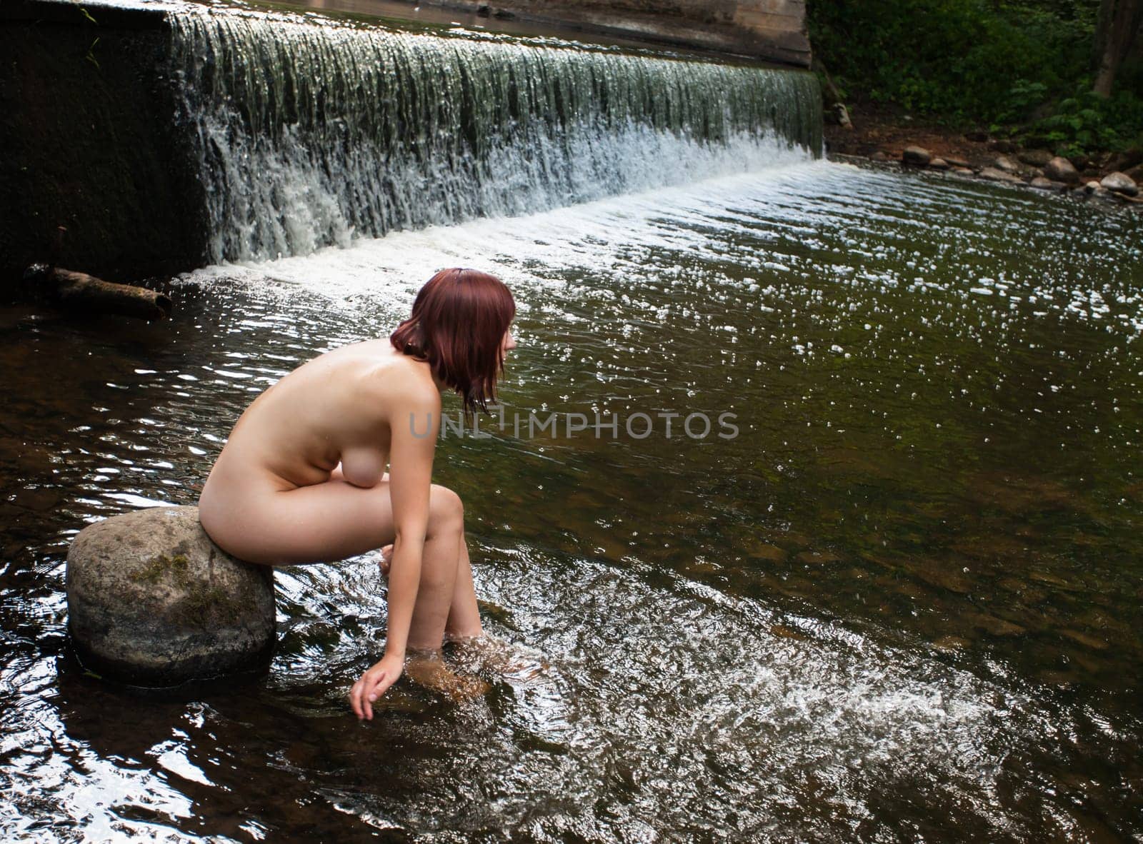 Young nude woman enjoying the fresh coolness near a forest stream with a waterfall