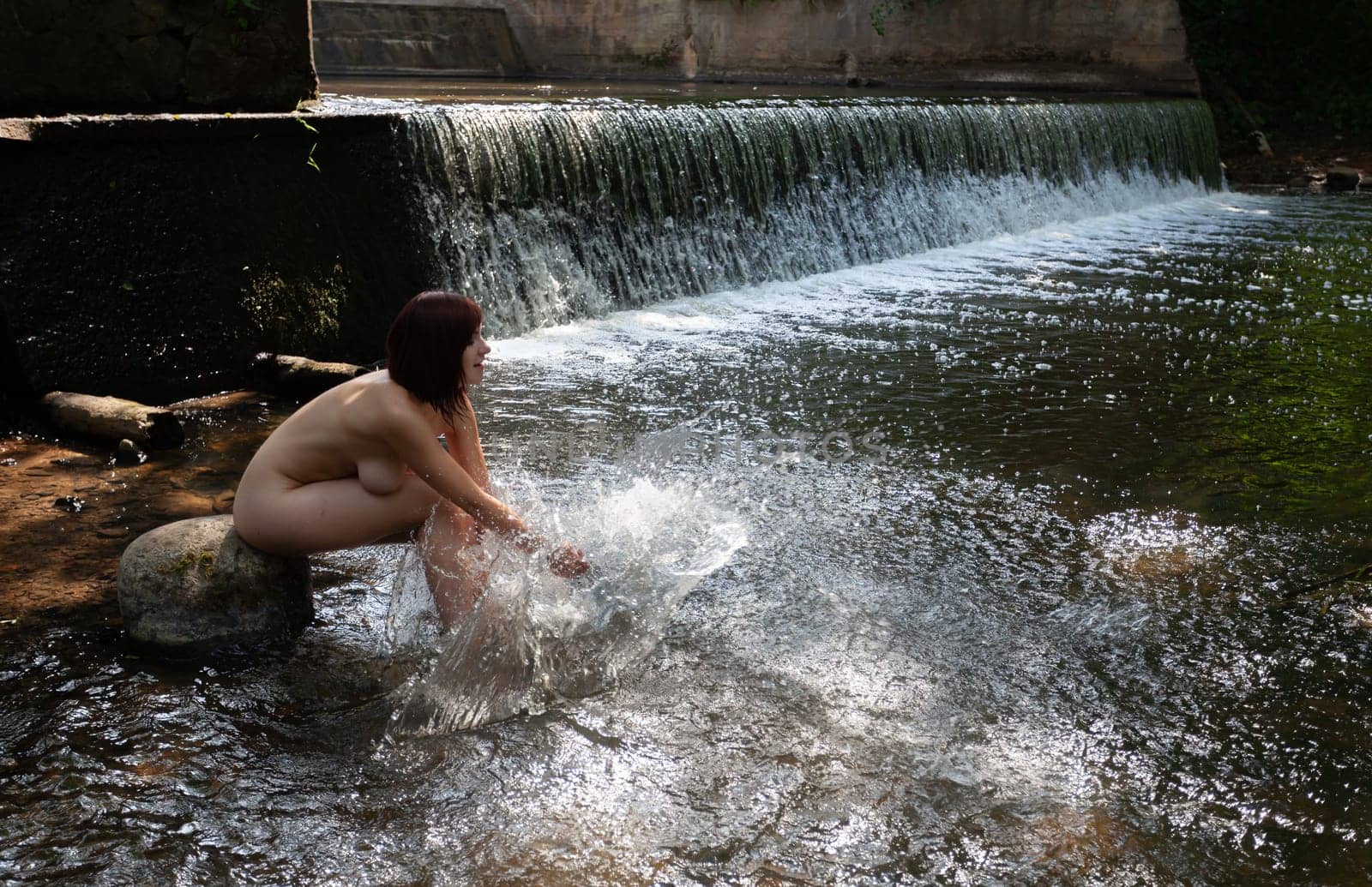 Young nude woman enjoying the fresh coolness near a forest stream with a waterfall