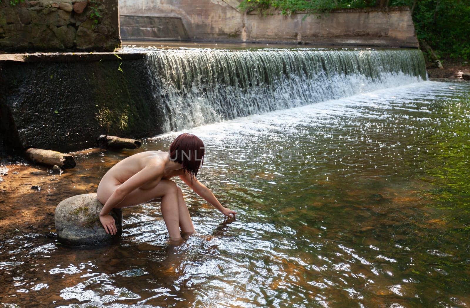 Young nude woman enjoying the fresh coolness near a forest stream with a waterfall