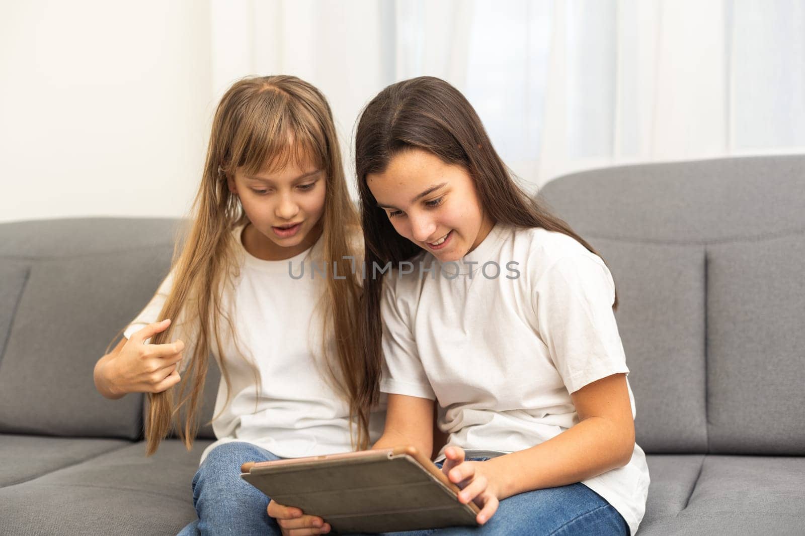 Young twins using a laptop and a tablet sitting on a couch in the living room. High quality photo