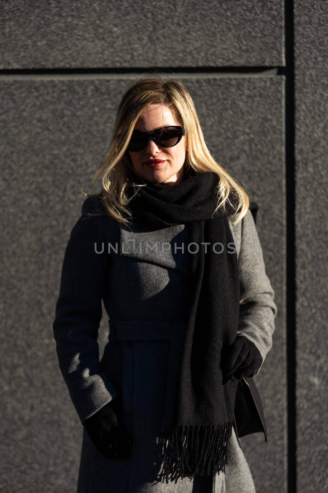 Blonde girl in sunglasses and a black coat against a wall. Urban portrait of a young woman. High quality photo