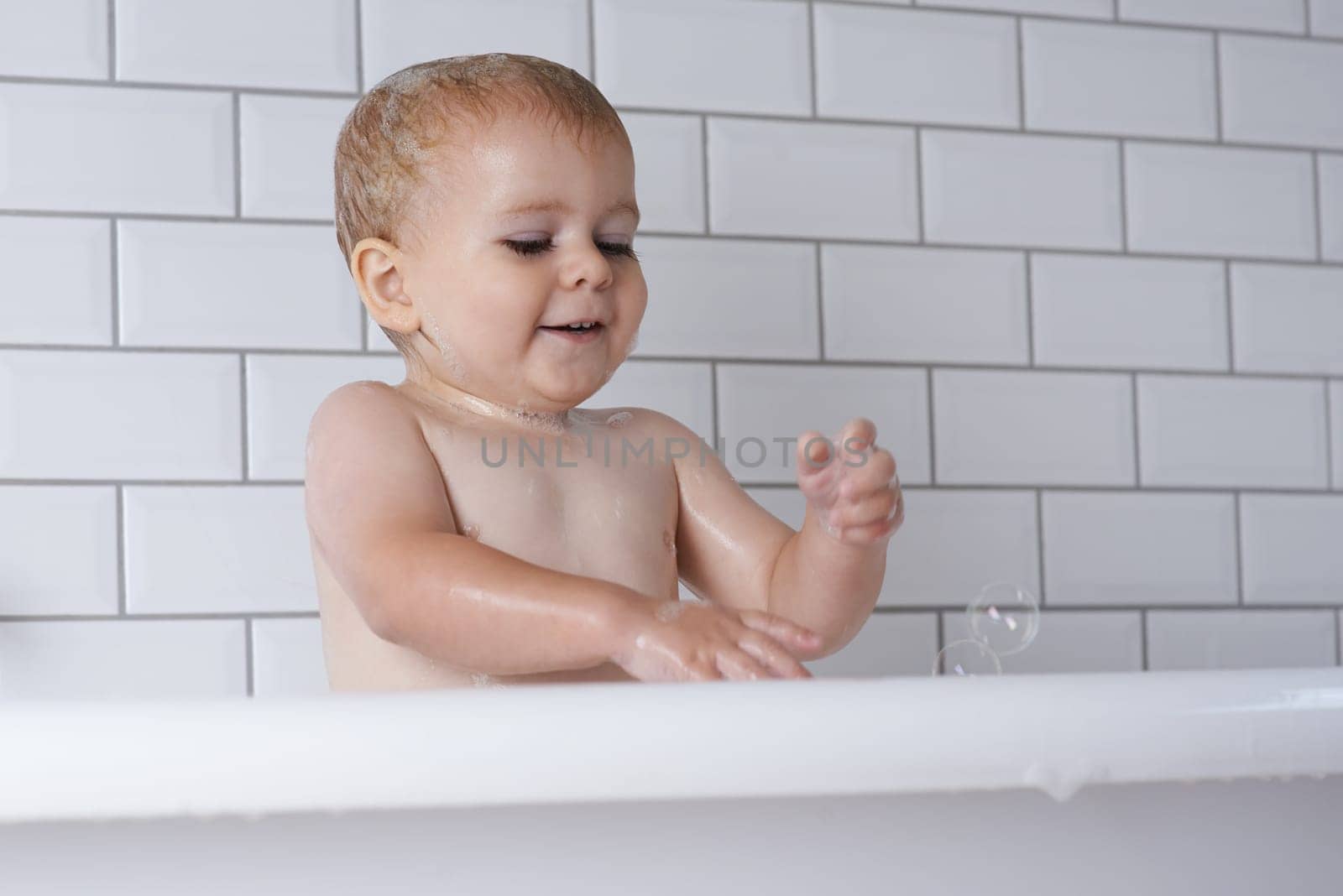 Baby, child and playing with bubbles in bath of water for morning routine, skincare and wellness at home. Happy boy, kid and cleaning in tub with soap, foam and washing for fun, hygiene and bathroom by YuriArcurs