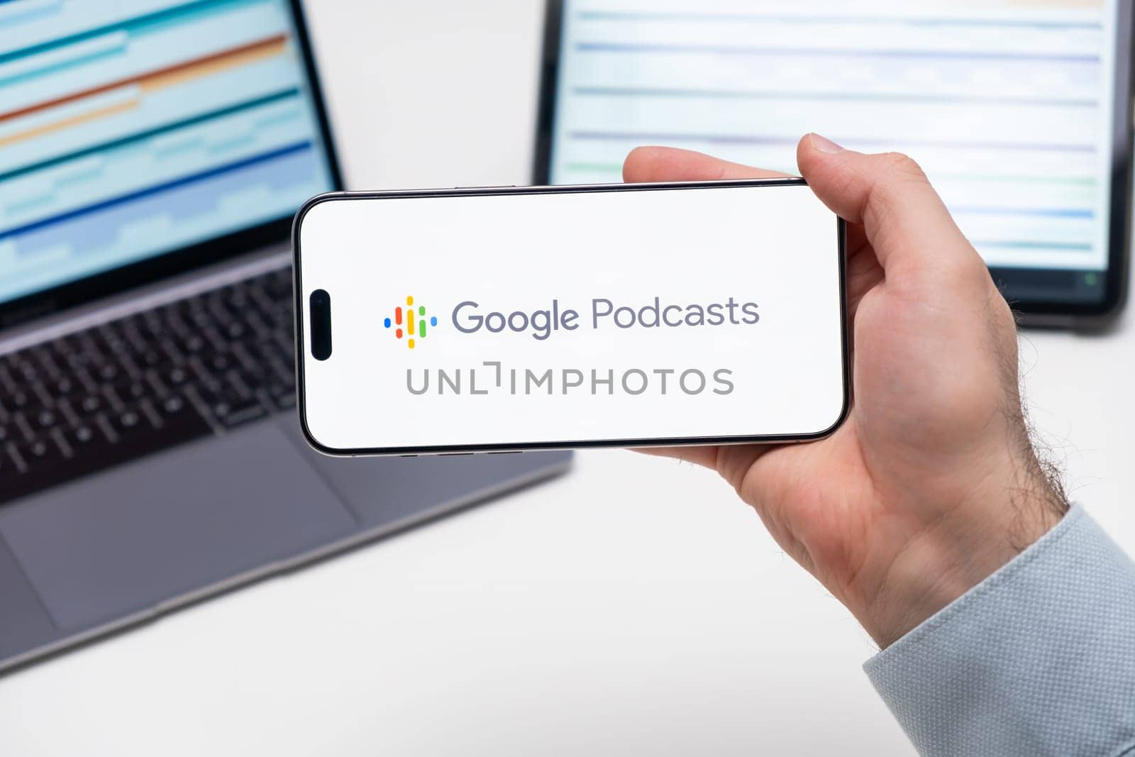 Google Podcast logo of app on the screen of mobile phone held by man in front of the laptop and tablet by vladimka