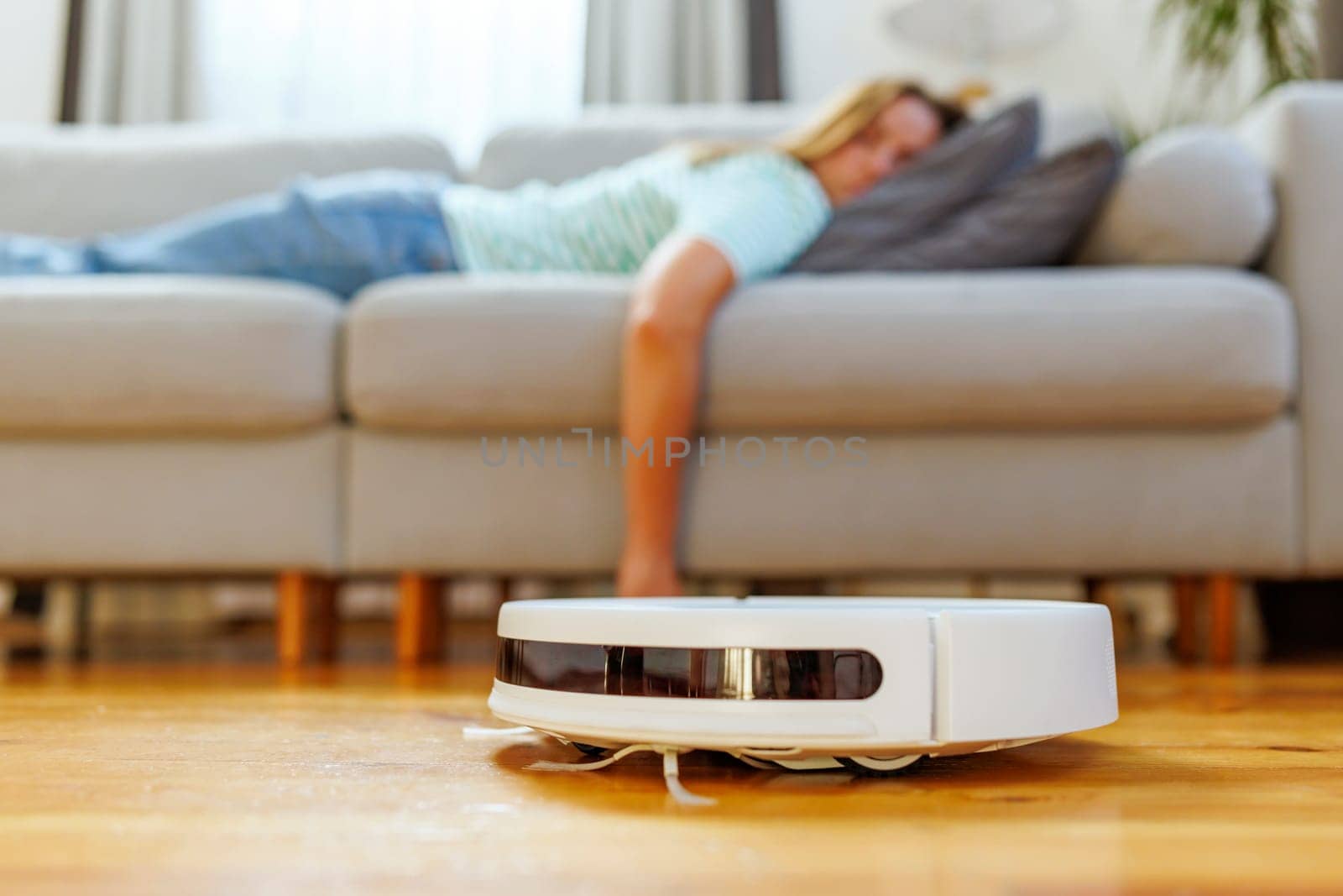 A woman takes a peaceful nap on the couch while the robotic vacuum cleaner quietly cleans the hardwood floor in a bright living room.