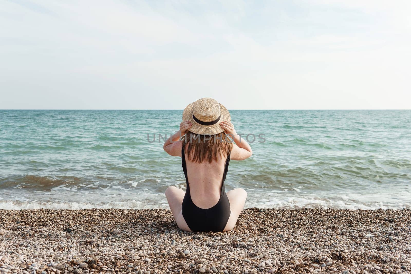 Woman in black swimsuit and straw hat on beach, seaside. View from the back. Summer vacation at the sea