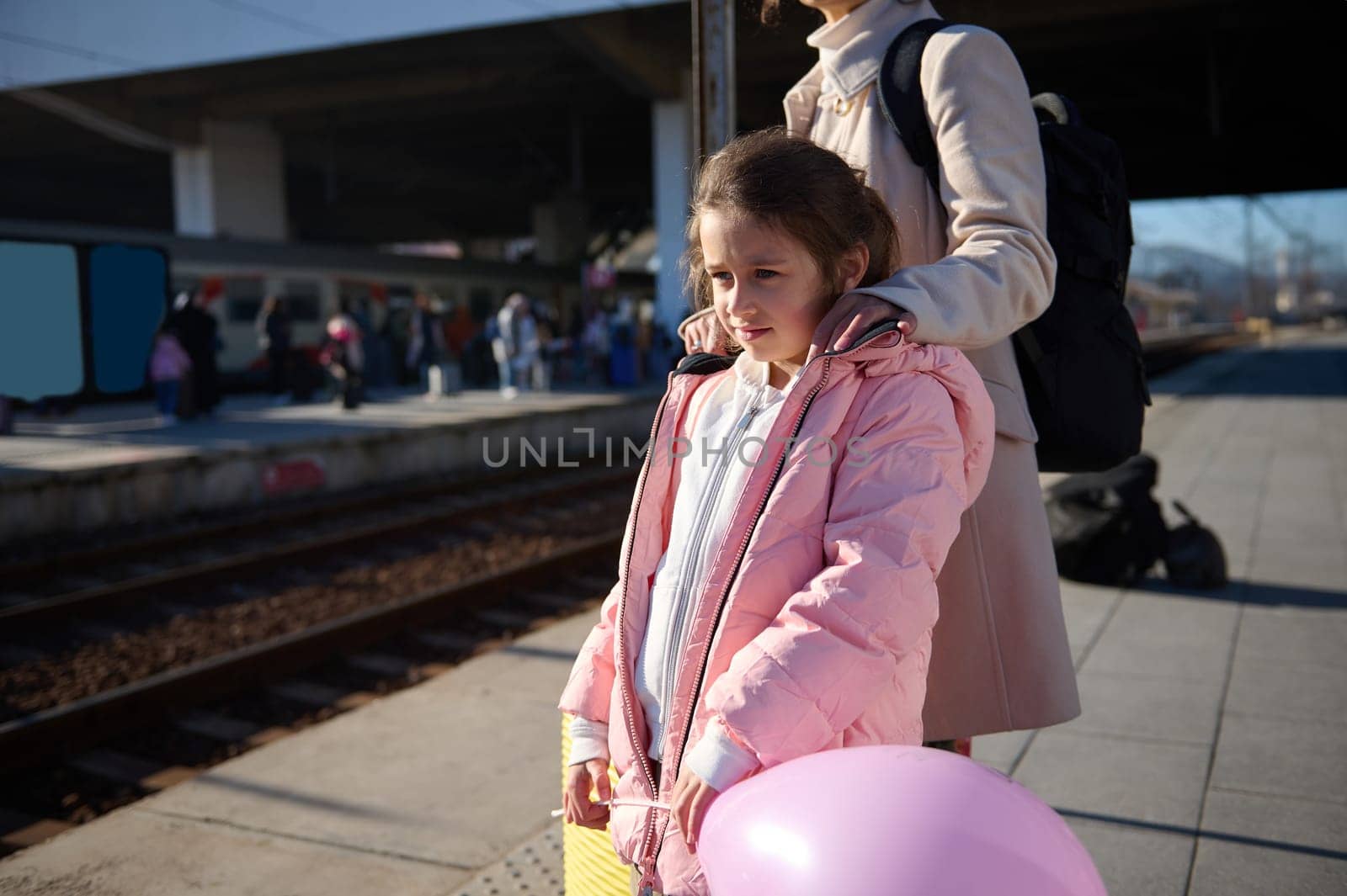Adorable Caucasian little child girl in warm pink down jacket, standing near her mom in the train station, waiting for train. Family travel by train. People. Trip. Lifestyle. Train as transportation