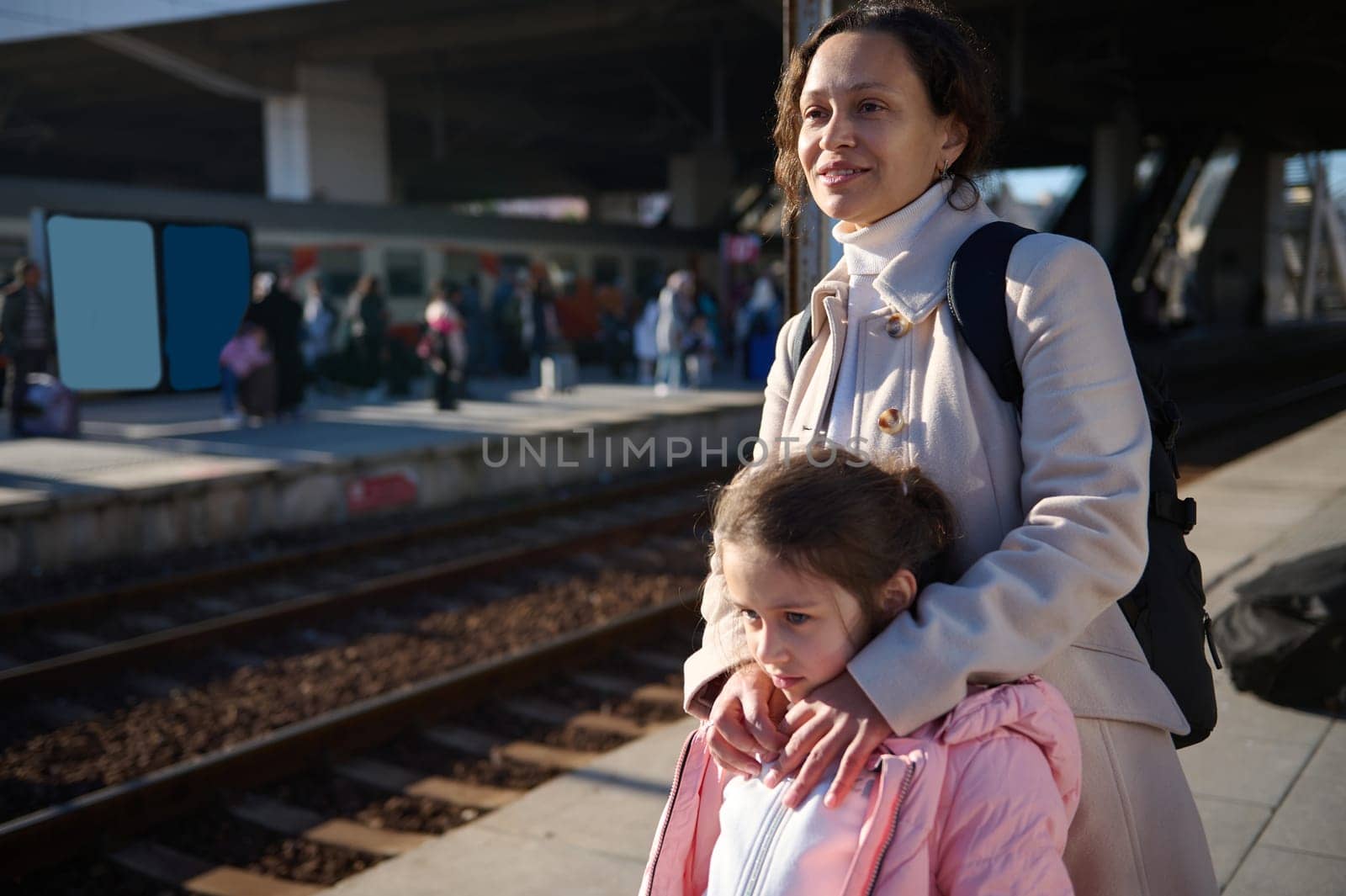 Young multiethnic woman, a mother and her little daughter waiting for the train, standing on the platform at the railway station.