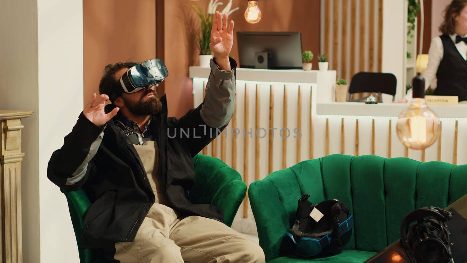 Traveler uses virtual reality technology in lounge area, having fun with vr interactive glasses while he wears winter jacket. Person having helmet and snowboard next to him, prepare to go skiing.