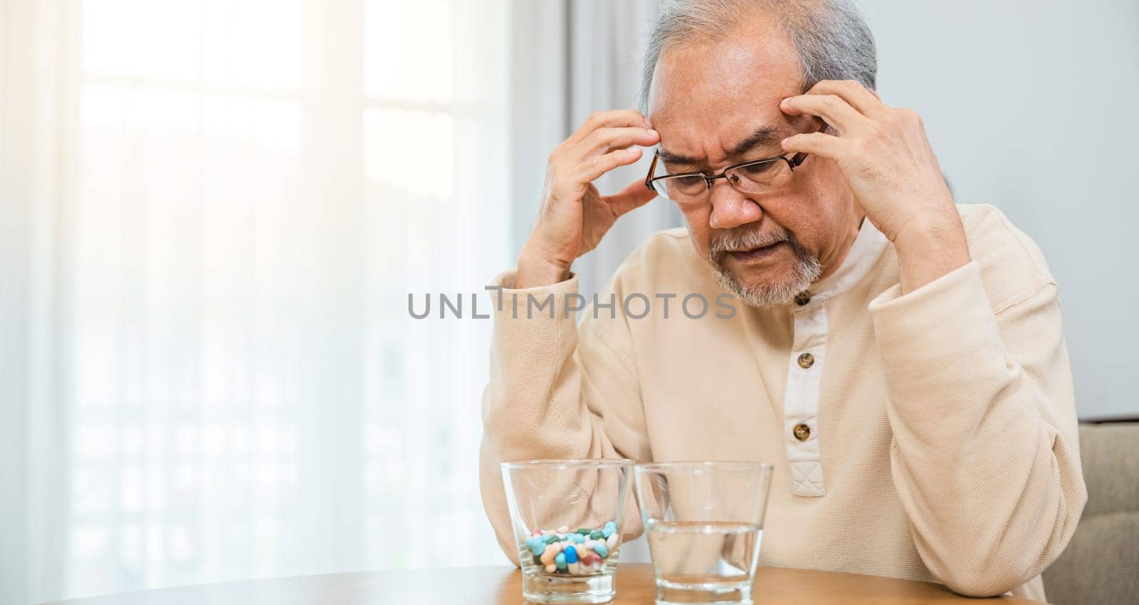 Asian senior old man grey haired sad sitting in chair hand hold head with many medicines and pills for taking, elderly man sick have headache looking pill glass on at home, Elderly healthcare