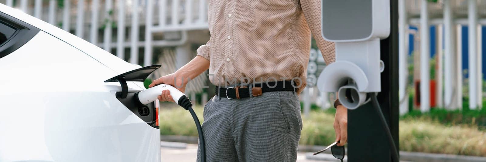 Young man recharge electric car's battery from charging station in city commercial parking lot. Rechargeable EV car for sustainable environmental friendly urban travel. Panorama Expedient