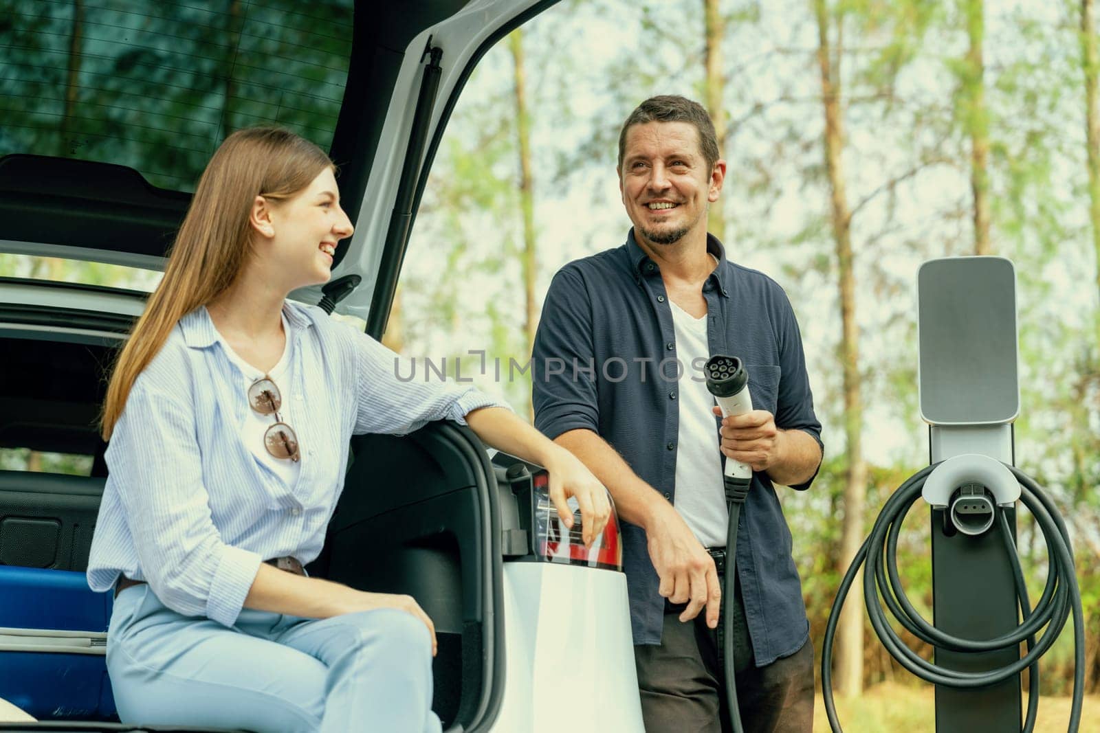 Lovely young couple recharging battery for electric car while relaxing and sitting on the trunk during road trip travel EV car in autumnal forest. Eco friendly travel on vacation during autumn. Exalt