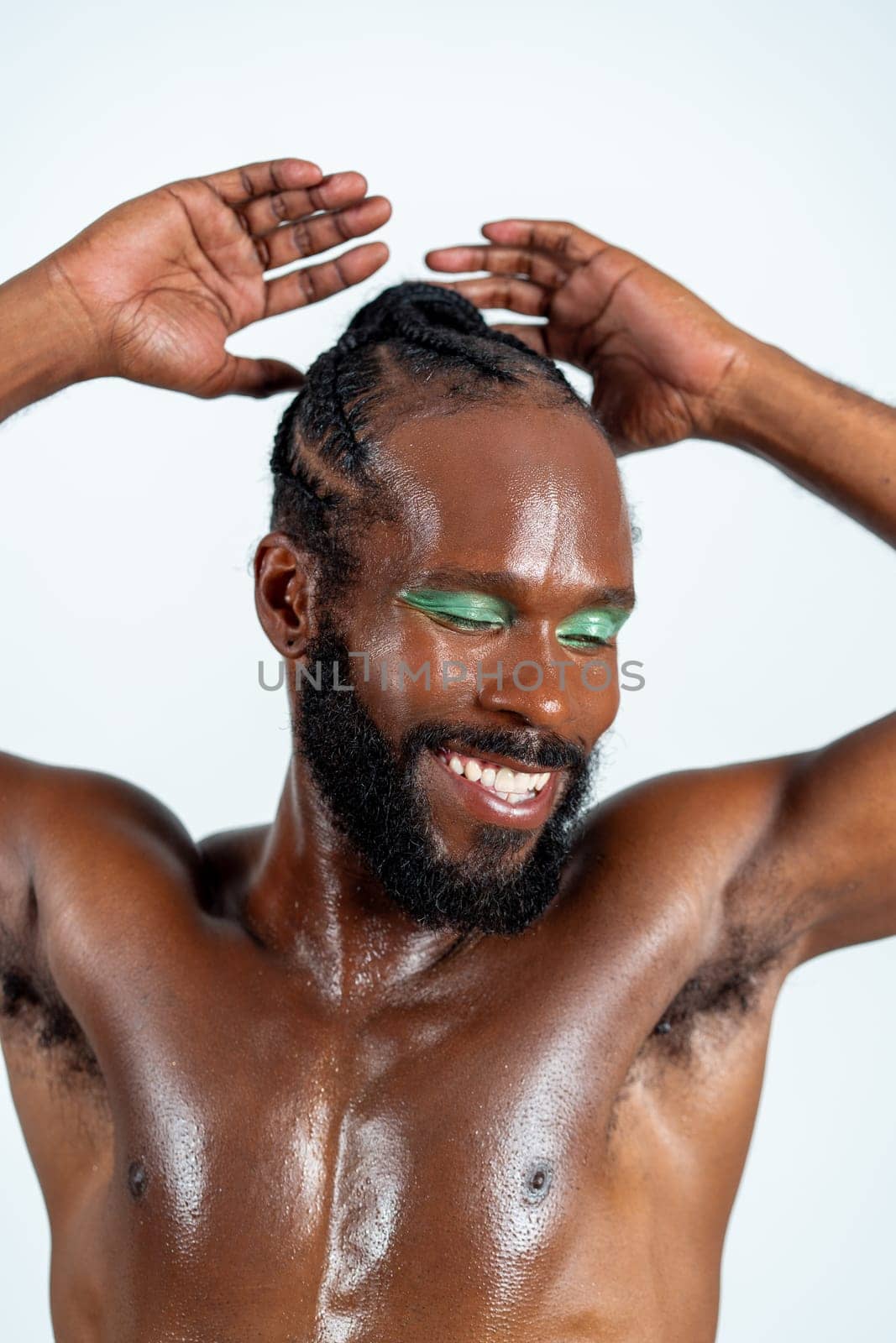 Portrait of shirtless muscular African American gay man with green eyeshadow. Confident bearded man smiling with hands raised on white background. Happy gay with bright makeup.