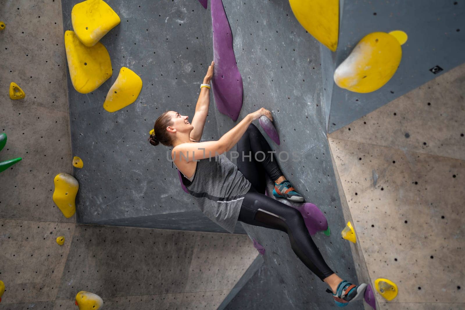 Side view of smiling sportswoman climbing artificial wall in gym. Full length of determined woman practicing bouldering in sports center. Health and fitness concept.