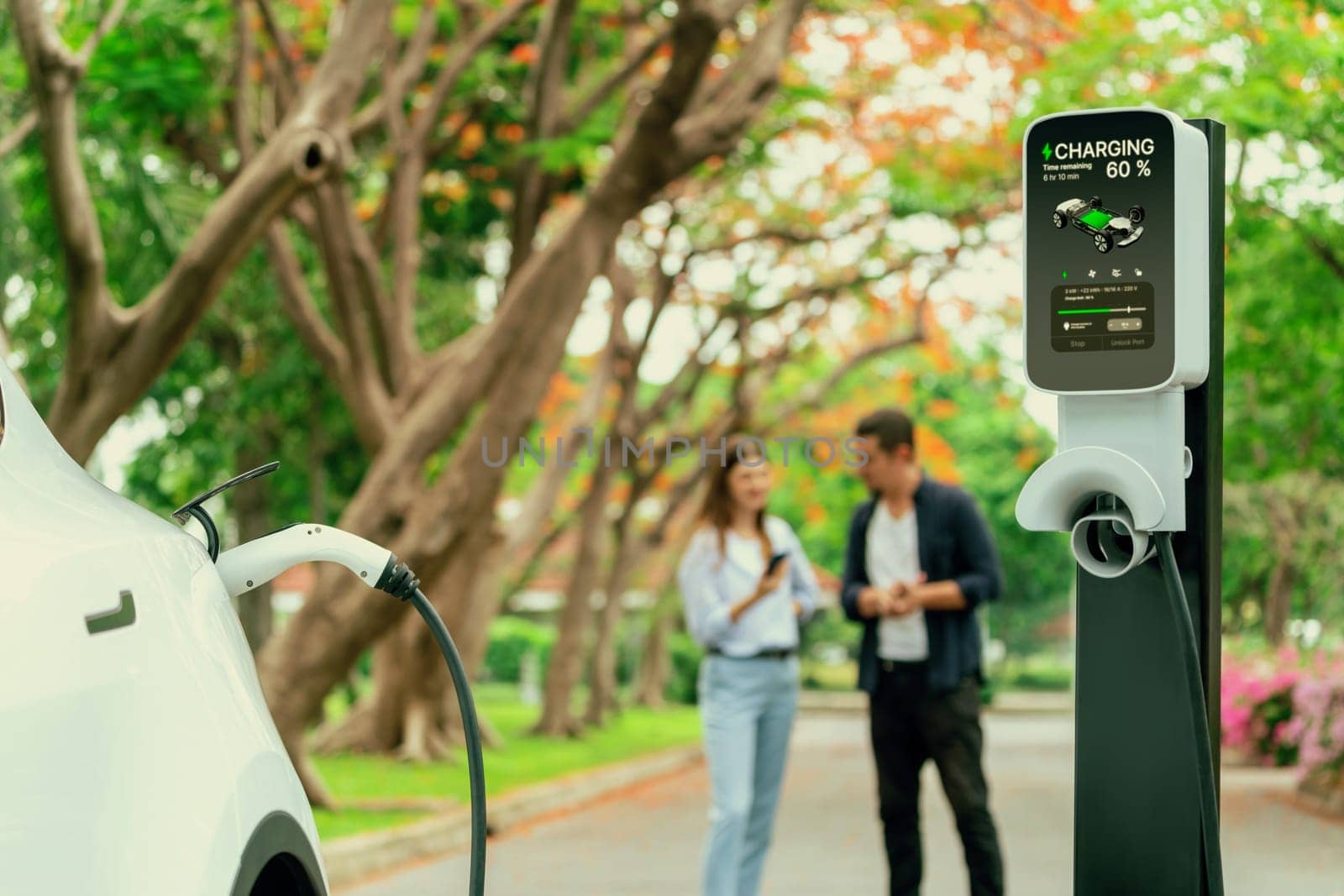 Focused EV car recharging battery on blurred background of lovey couple during autumnal road trip travel with electric vehicle recharging battery. Eco friendly travel on vacation during autumn. Exalt