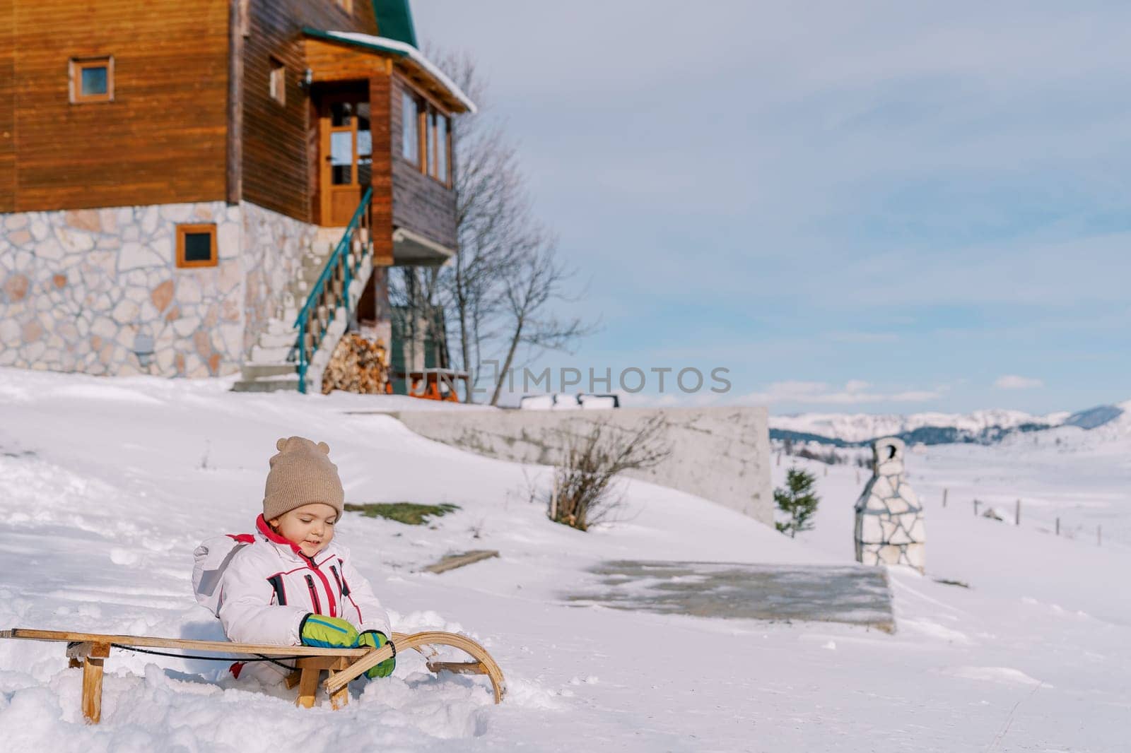 Little girl sits in the snow near a wooden sled in the courtyard of a house. High quality photo