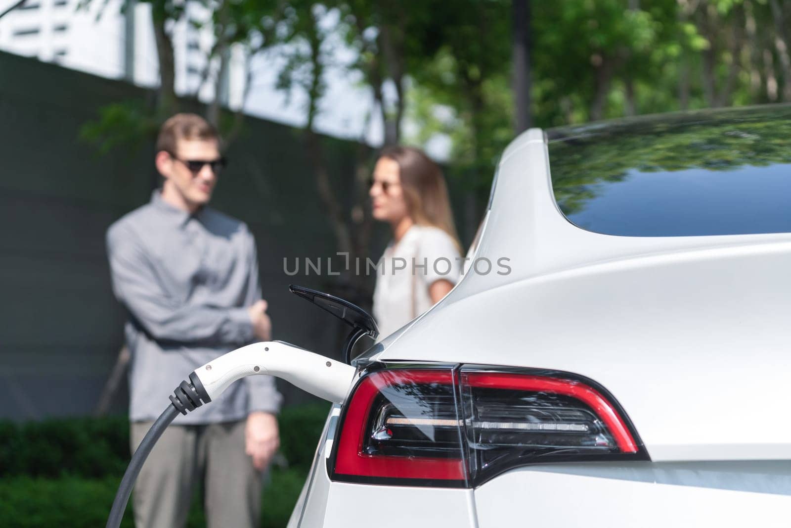 Young couple travel with EV electric car charging in green sustainable city outdoor garden in summer shows urban sustainability lifestyle by green clean rechargeable energy of electric vehicle innards