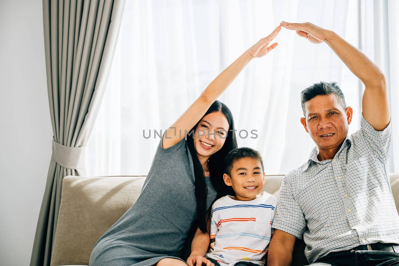 A joyful family poses on a sofa parents making a roof gesture above their little son by Sorapop