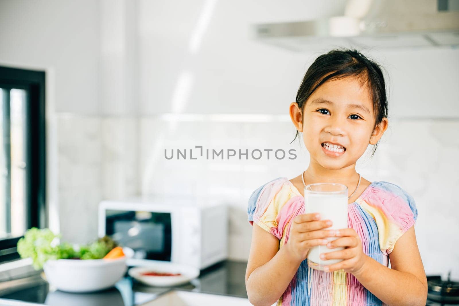 Young Asian girl in kitchen holds milk smiling cheerfully. Portrait of cute daughter enjoying drink radiating joy. Happy preschooler sips calcium-rich liquid feeling cheerful at home give me. by Sorapop