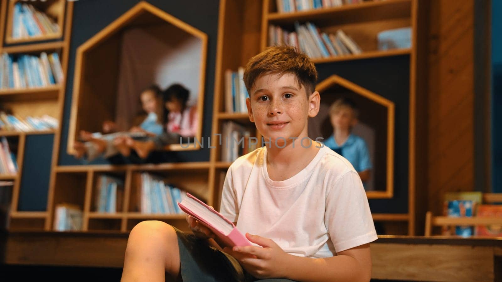 Caucasian boy looking at camera while group of smart students sitting at library. Child studying, learning, reading from novel or textbook while children talking, chitchat about education. Erudition.