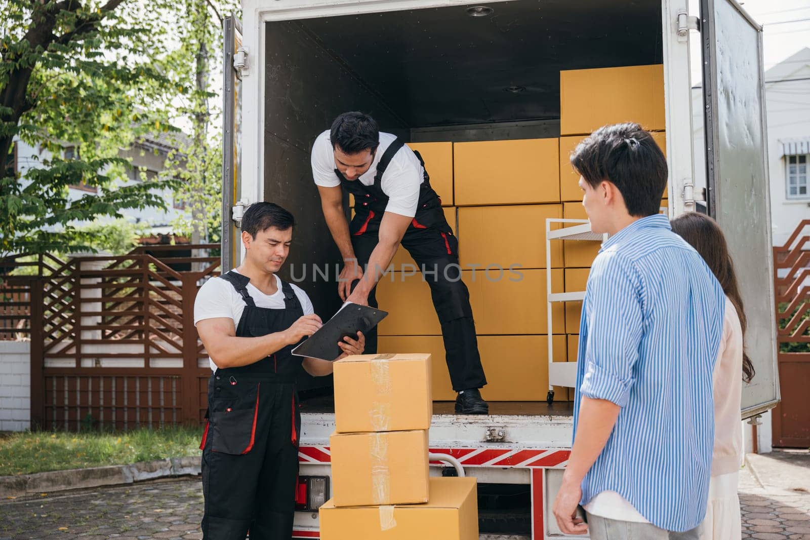 Professional movers aid a satisfied couple who signs the delivery checklist after furniture upload. Teamwork and customer satisfaction are evident. Moving Day Concept by Sorapop