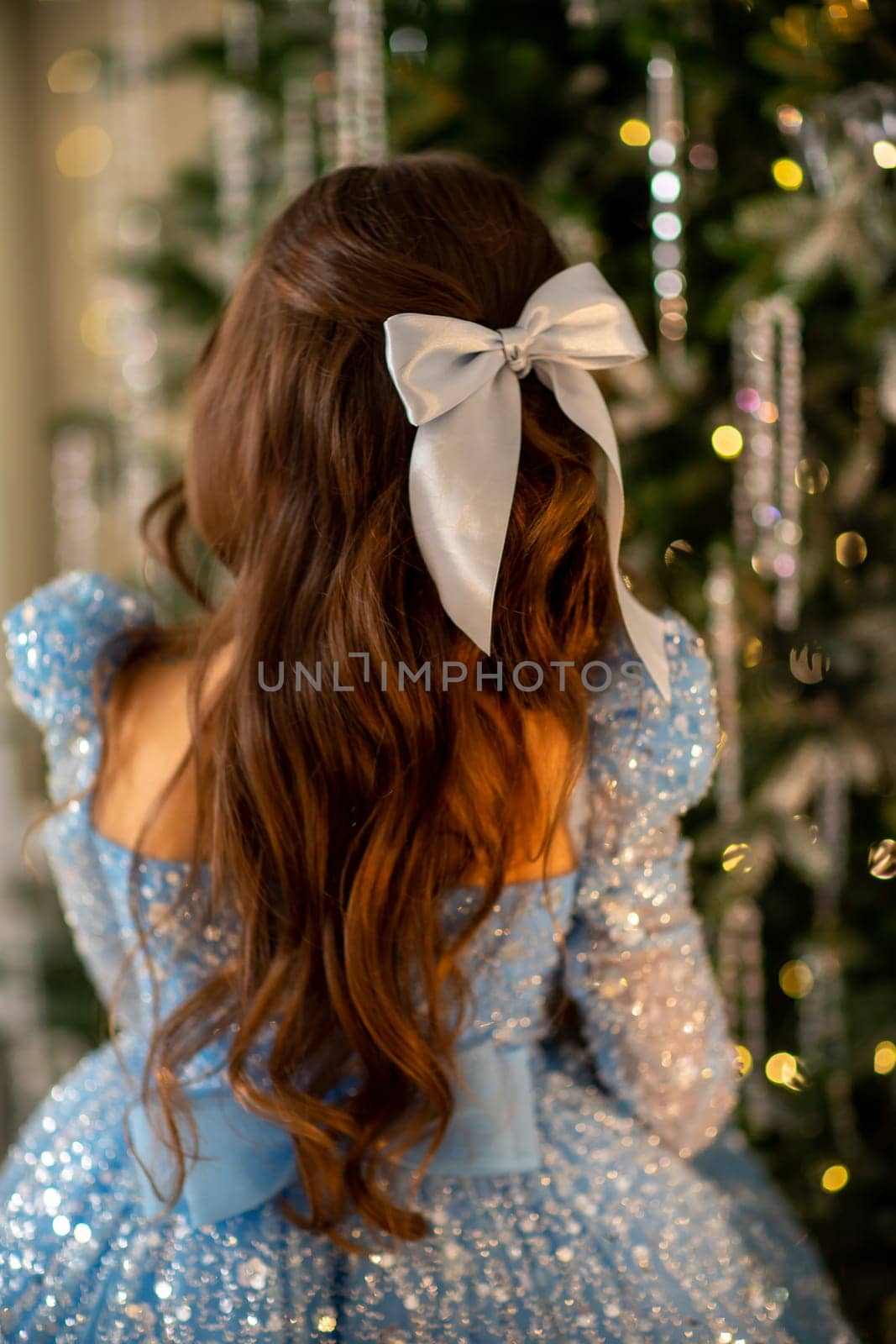The girl decorates the Christmas tree, holding Christmas baubles in her hand. She wears a blue dress with a bow in her hair. Holiday concept