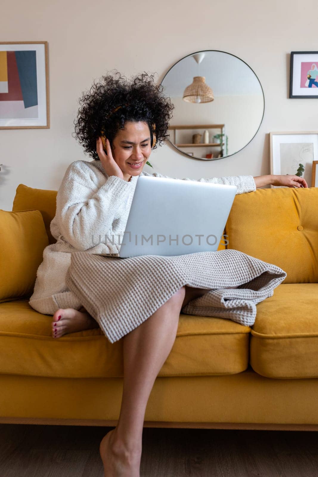 Happy African american woman watching movie with laptop and headphones sitting on the couch relaxing at home. Vertical. by Hoverstock