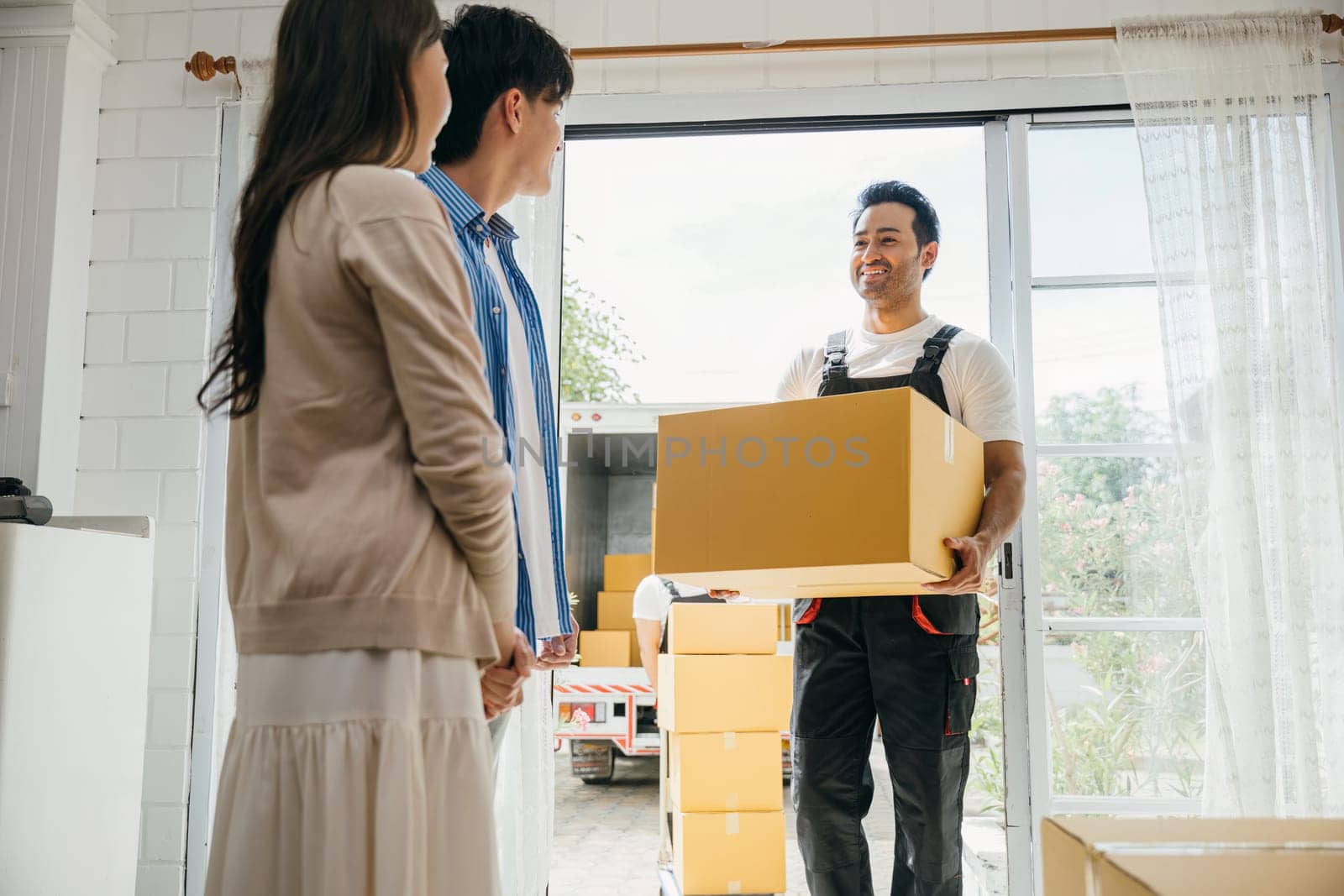 Couple receives moving assistance inside home. Movers in uniform unload boxes from truck ensuring smooth relocation. Professional service teamwork and customer satisfaction shown. Moving new house