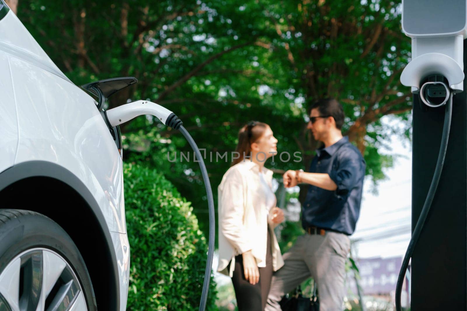 Young couple recharge electric car's battery from charging station in outdoor green city park in springtime. Rechargeable EV car for sustainable environmental friendly urban travel lifestyle.Expedient