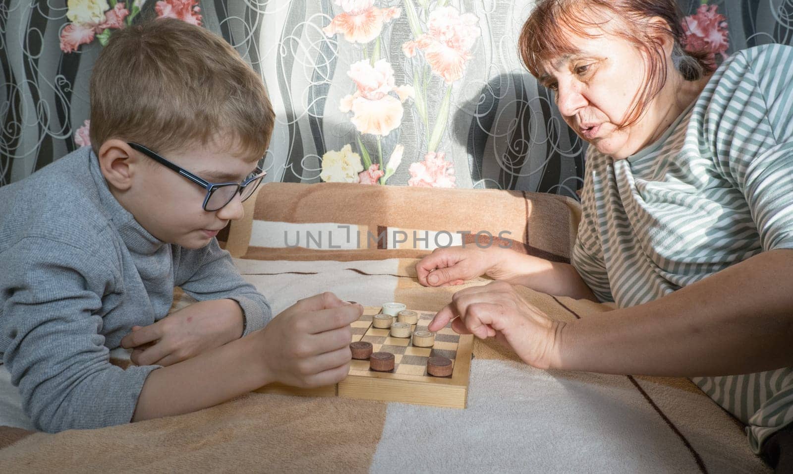 A caring middle-aged grandmother plays checkers with a child, a boy, lying on the bed. Happy family enjoying an interesting wooden board game at home