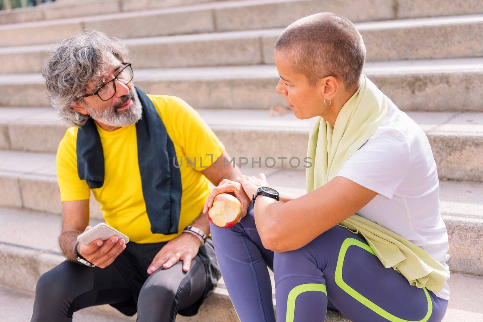 senior sportsman resting after training talking with his personal trainer who is eating an apple, concept of active and healthy lifestyle