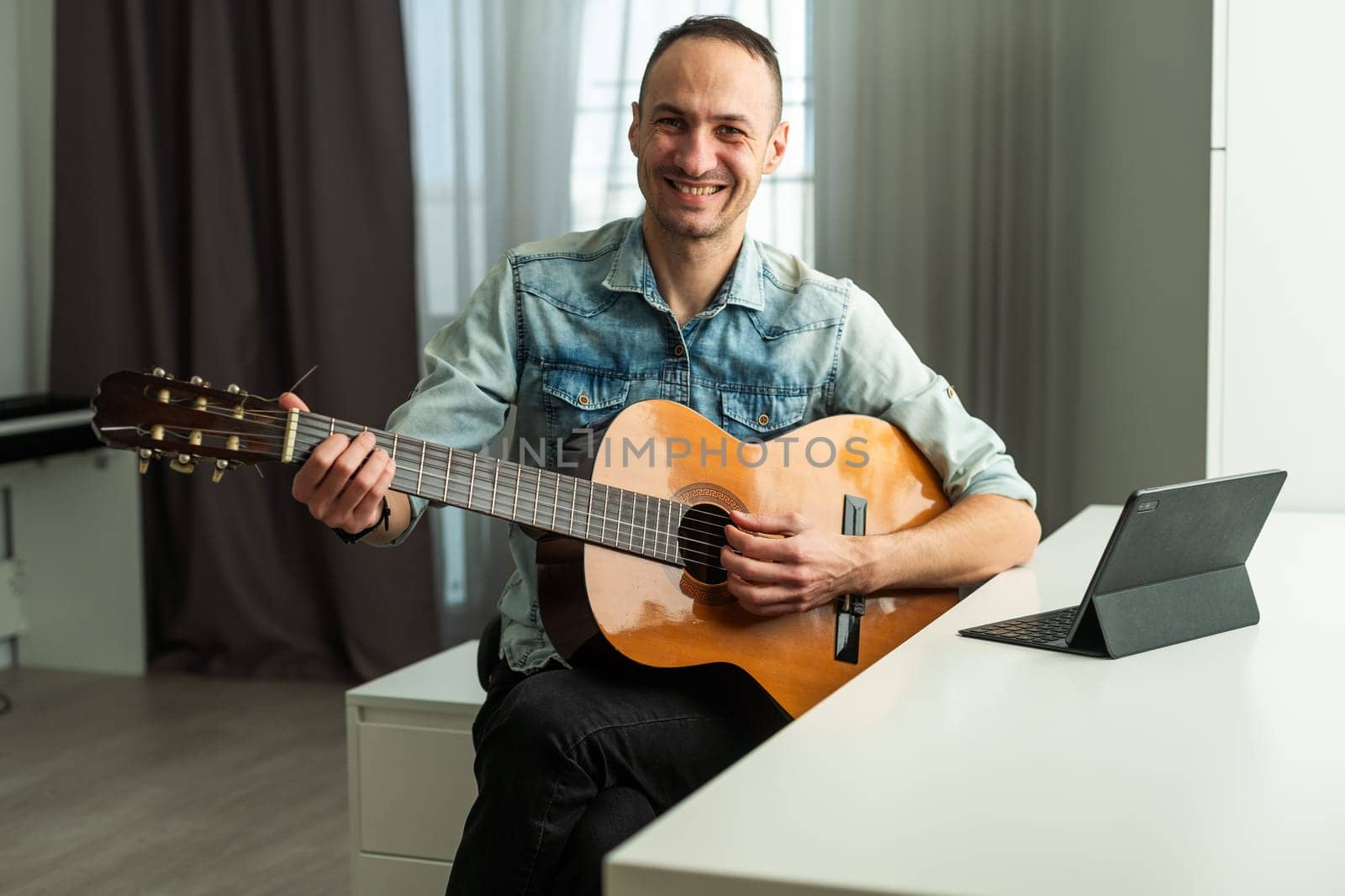 Portrait of man taking online guitar lesson looking at laptop screen. Retired male learning to play guitar watching webinar on computer at home by Andelov13