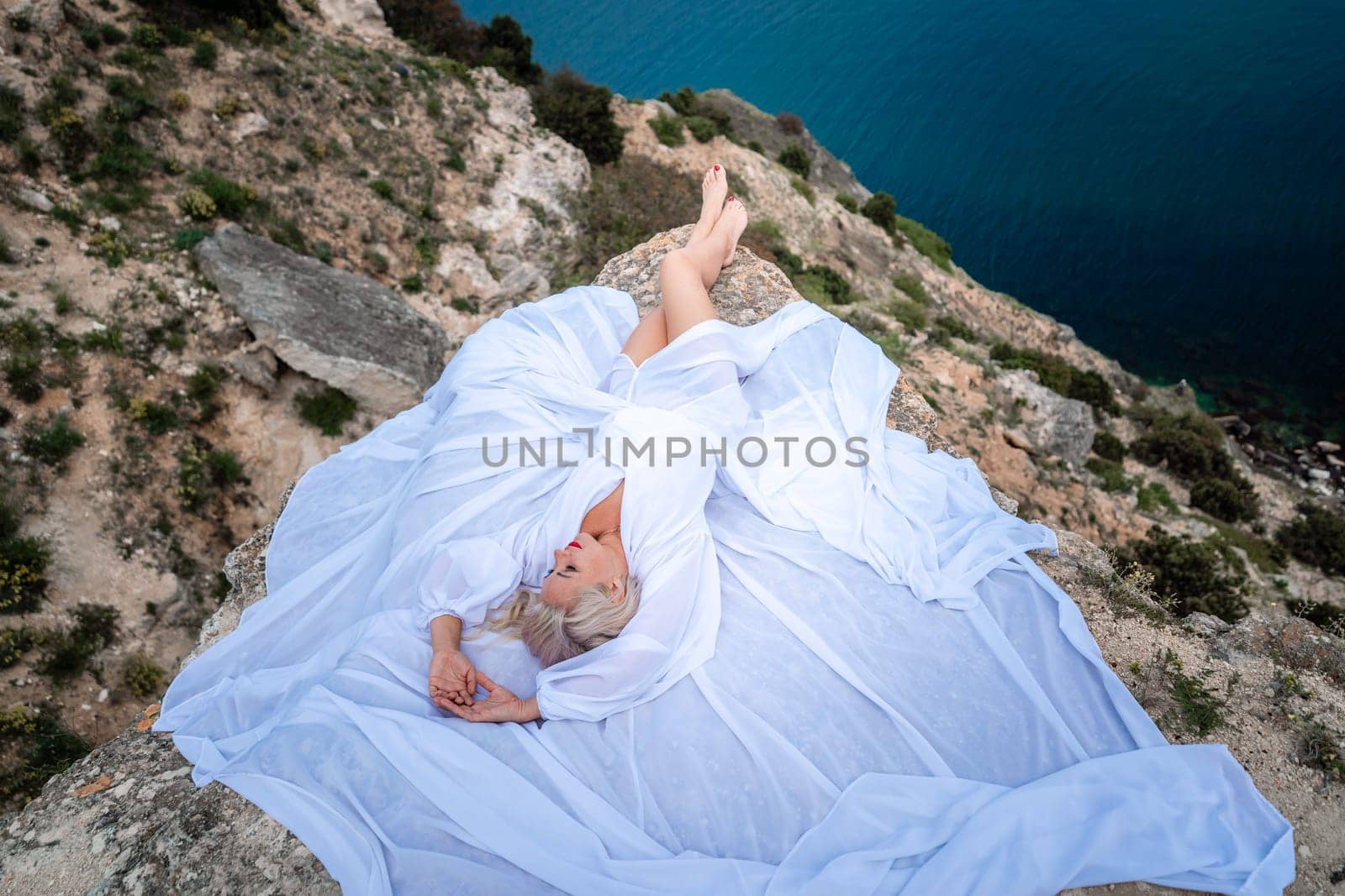 The blonde lies on a rock above the sea. With long hair on a sunny seashore in a white dress, rear view, silk fabric flutters in the wind. Against the backdrop of blue skies and mountains. by Matiunina