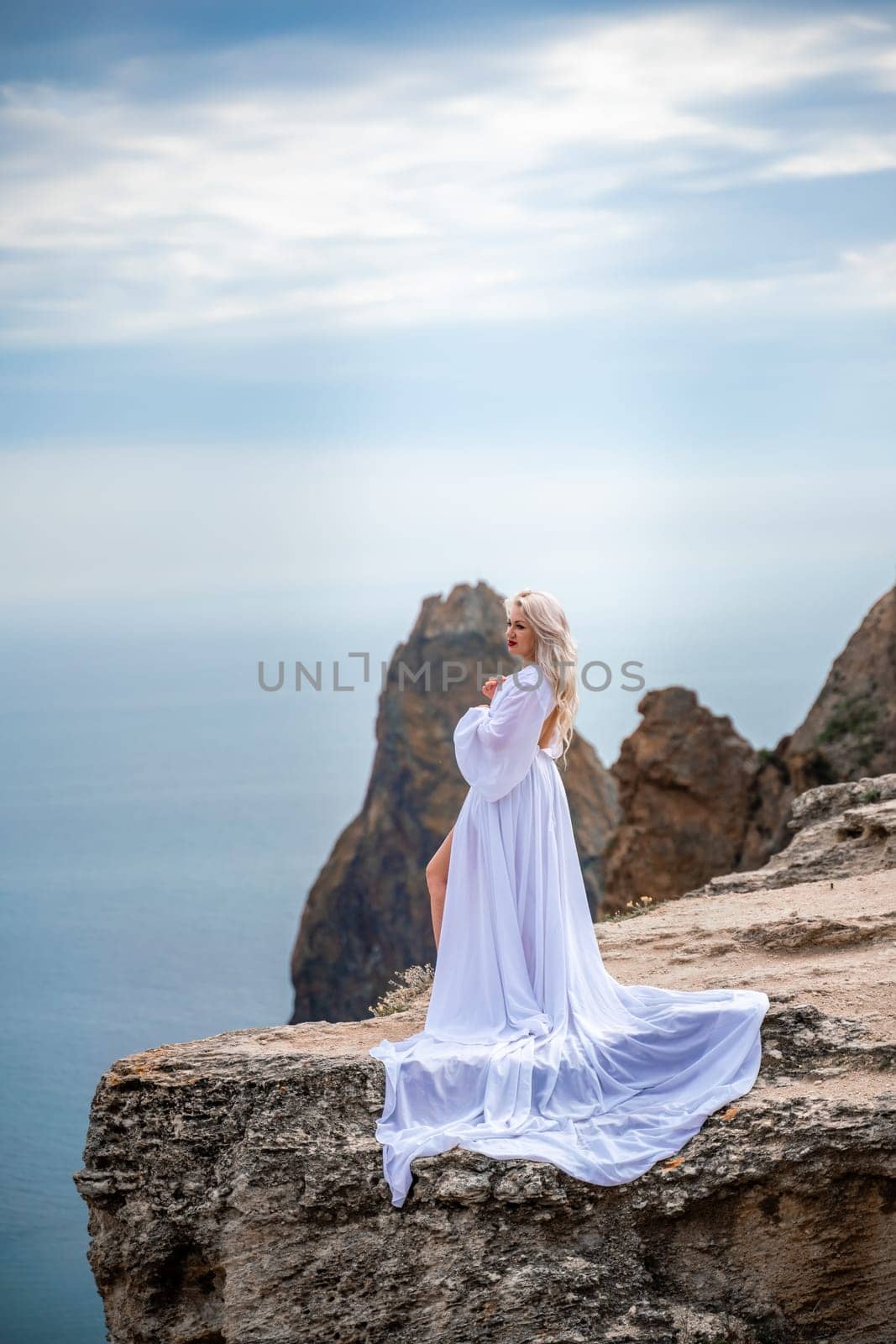 Blonde with long hair on a sunny seashore in a white flowing dress, rear view, silk fabric waving in the wind. Against the backdrop of the blue sky and mountains on the seashore