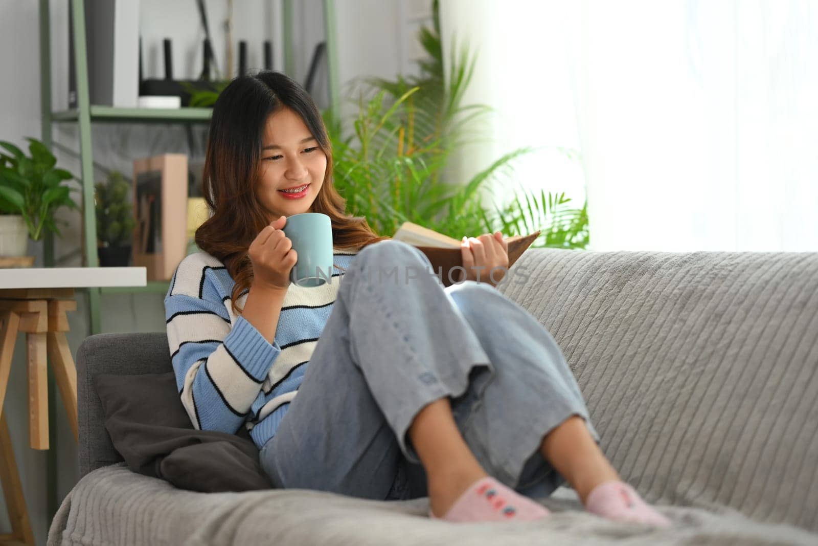 Relaxed young woman reading book and drinking coffee on couch at home by prathanchorruangsak